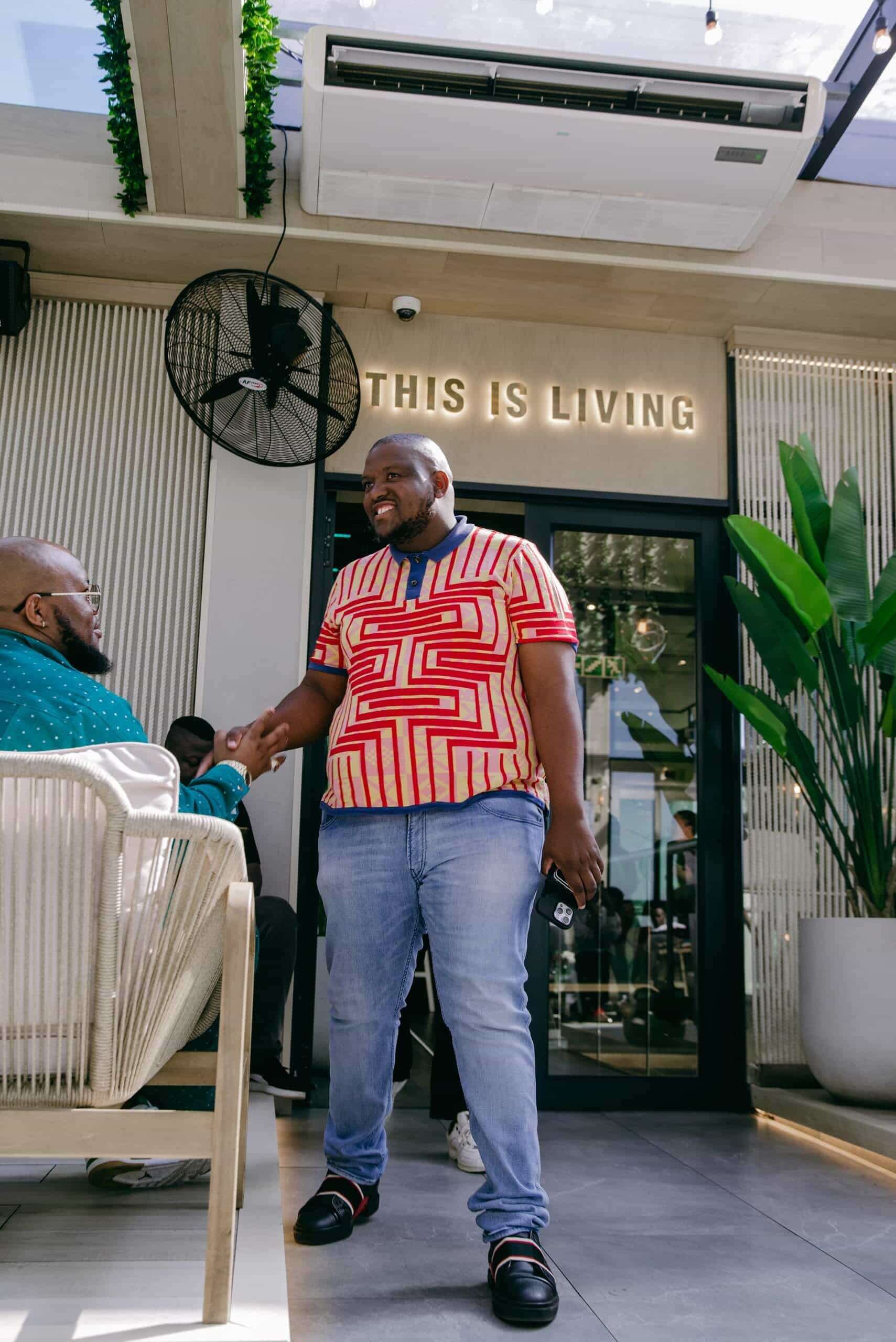 A smiling man in a red and yellow patterned shirt and jeans shakes hands with a seated person in a modern, sunlit indoor space. Large green plants and a sign reading &ldquo;THIS IS LIVING&rdquo; are visible above the glass door behind him.