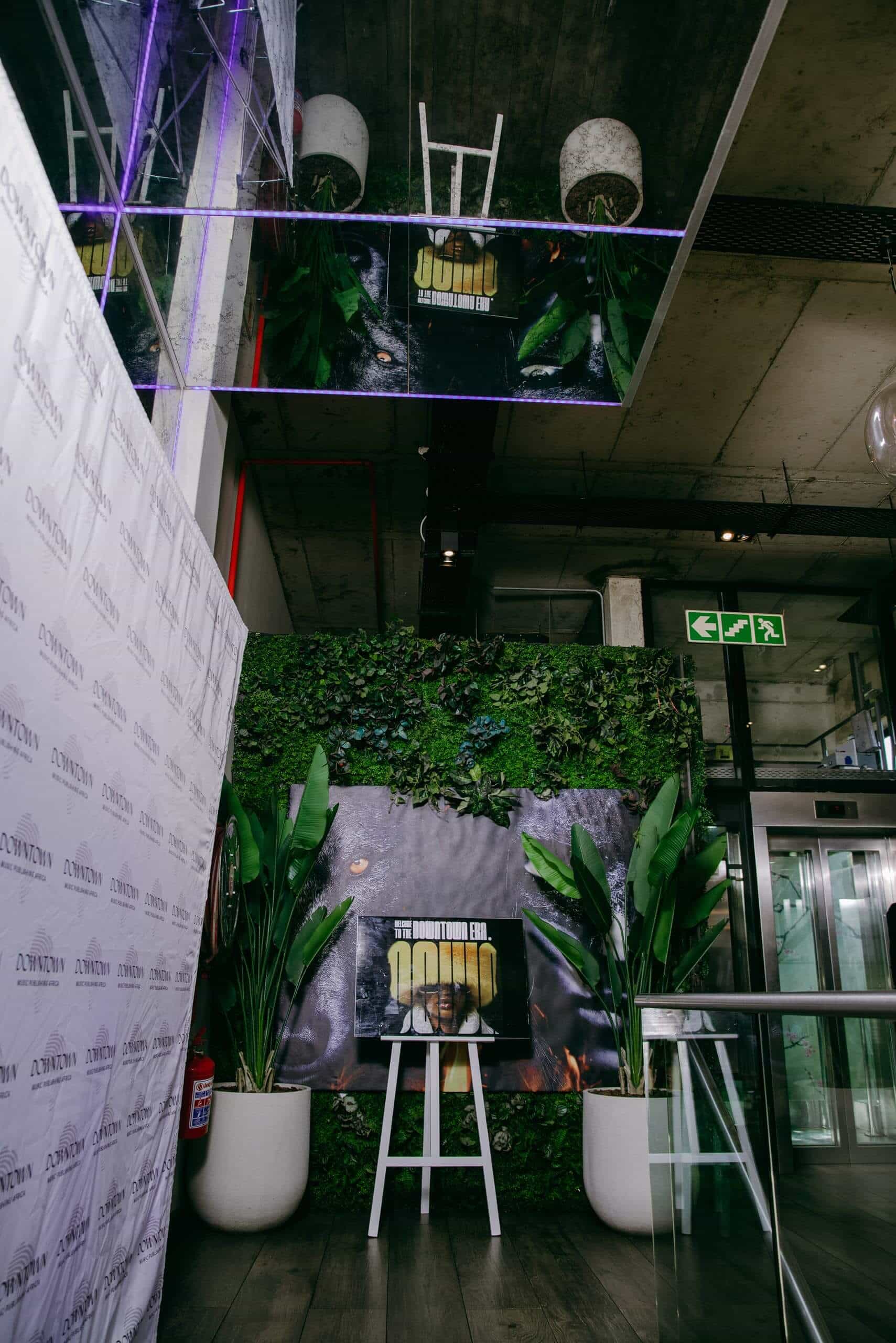 A modern indoor space with a green plant wall, large potted plants, a mirror on the ceiling, and a promotional sign on an easel that says 