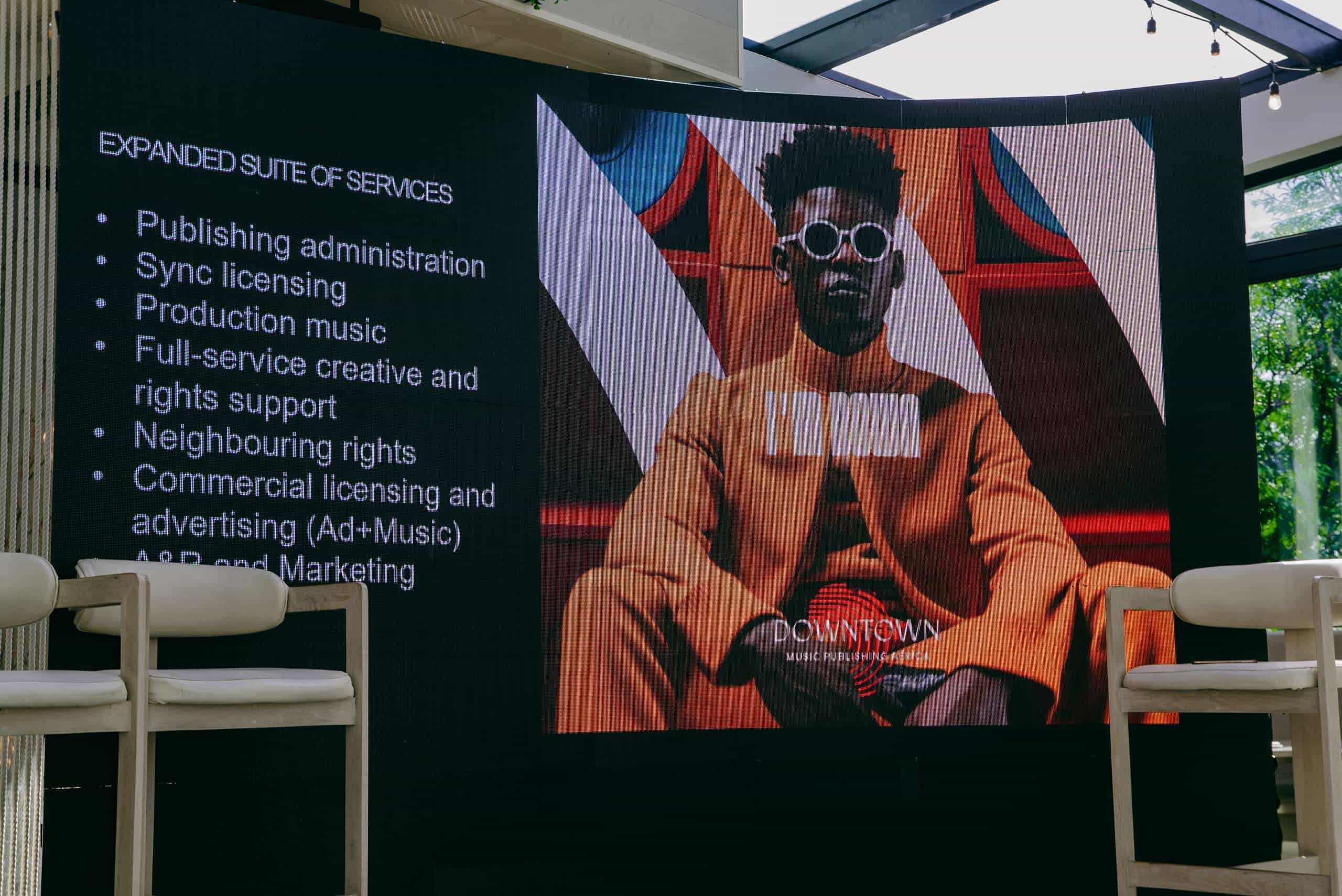 A large screen displays a music industry presentation. The left side lists services like publishing administration and sync licensing. The right side shows an album cover featuring a stylish person in sunglasses and orange clothes, with 