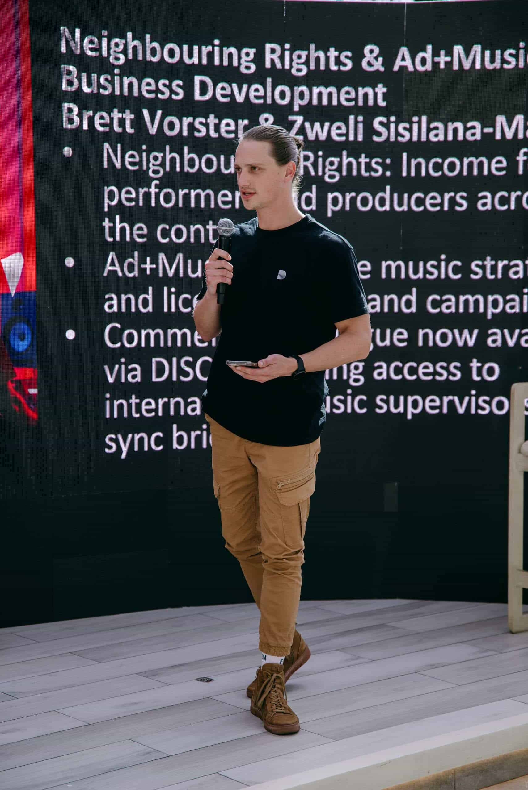 A person in a black t-shirt, tan cargo pants, and brown shoes holds a microphone and phone, standing on a light wood floor. Behind them is a large screen with white text about music business development and neighbouring rights.