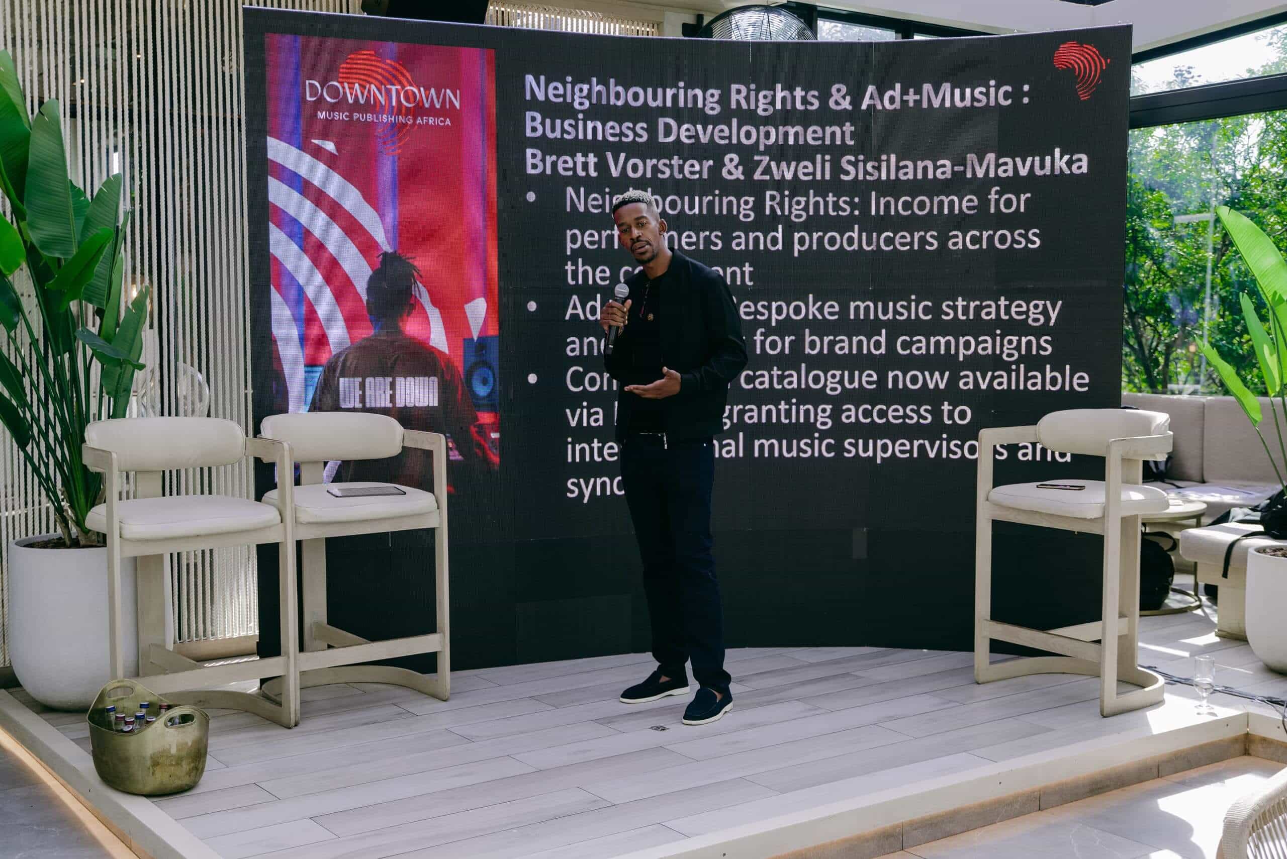 A man stands on stage with a microphone in front of a large screen displaying a music industry presentation. Plants, chairs, and sunlight fill the modern indoor space. The slide discusses 