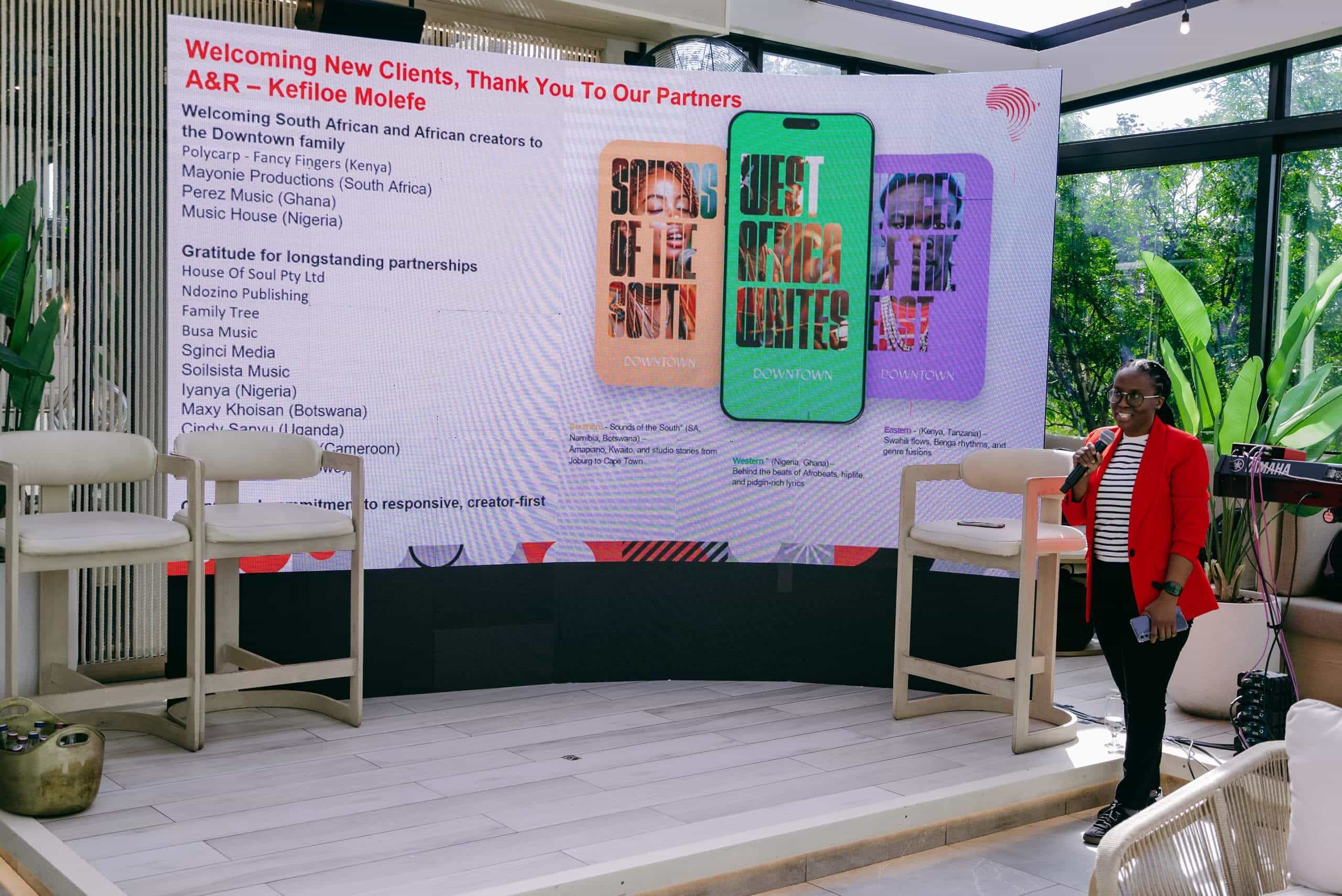 A woman in a red blazer stands by a large screen displaying colorful graphics of two smartphones and text about welcoming new clients and partners. The indoor space has natural light, green plants, and white chairs arranged on a wooden floor.