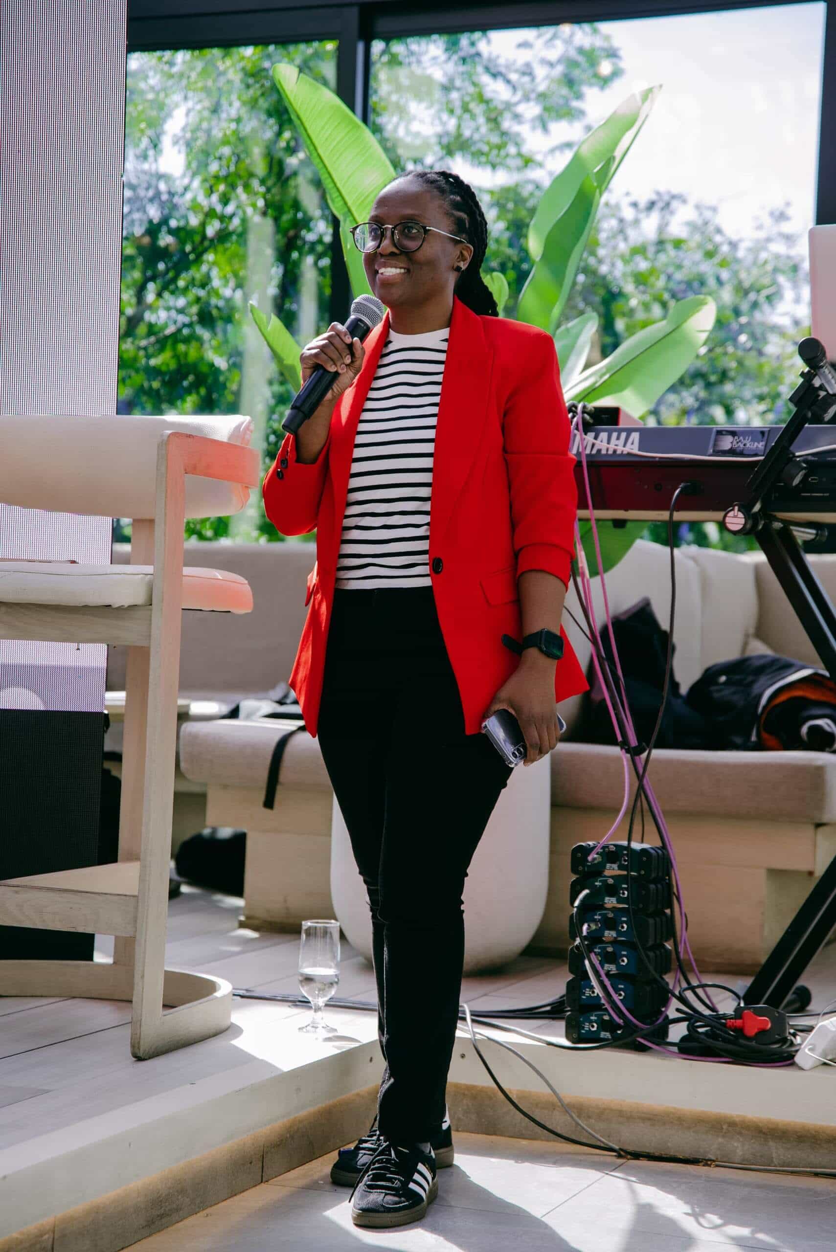 A smiling person wearing glasses, a red blazer, black-and-white striped shirt, black pants, and sneakers holds a microphone while standing indoors by a keyboard and audio equipment. Sunlight streams through large windows with leafy green plants outside.