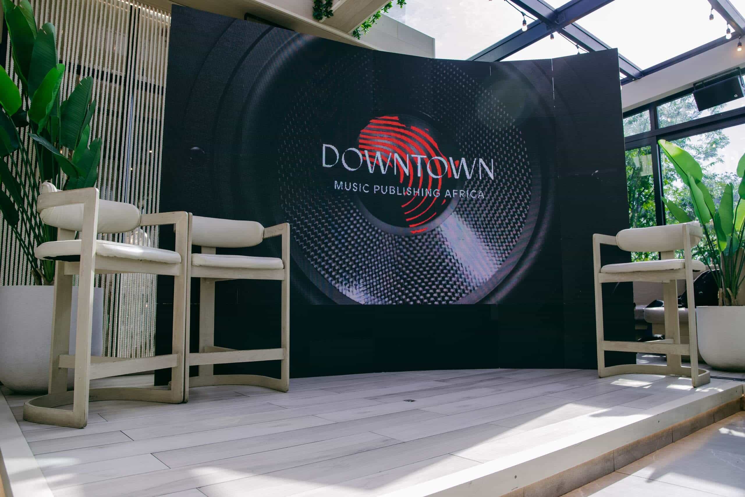 A stage with two beige chairs faces a large curved screen displaying the Downtown Music Publishing Africa logo with a red fingerprint. The area is surrounded by tall green plants and natural light enters through large windows above.