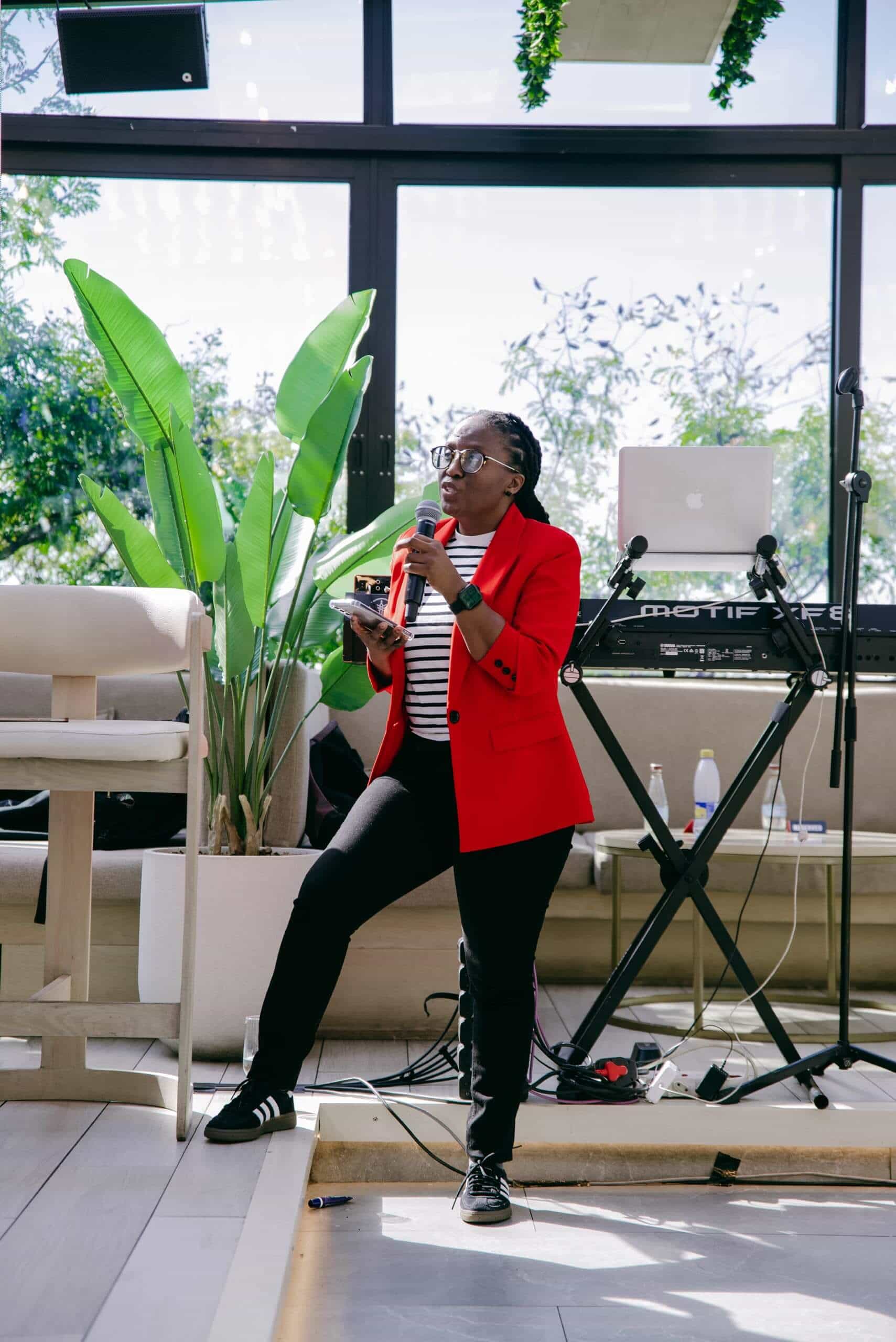 A person in a red blazer, striped shirt, and black pants speaks into a microphone, holding a phone. They stand on a stage with one leg resting on a step. Behind them are a keyboard, microphone stand, potted plant, and large windows showing greenery outside.