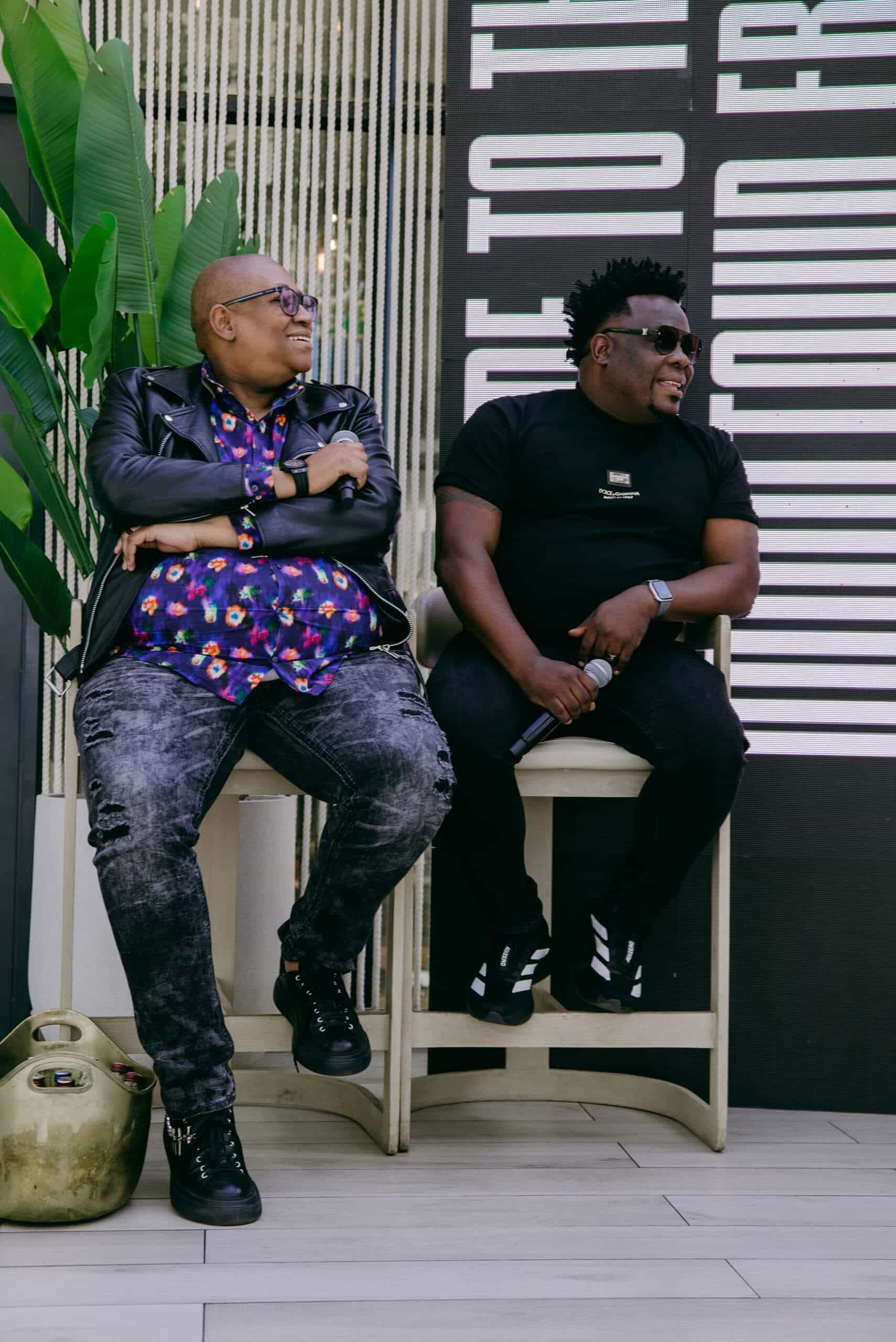 Two people sit casually on stools, smiling and chatting. One wears a black leather jacket over a colorful shirt and patterned jeans; the other wears a black t-shirt, pants, and sunglasses. They both hold microphones. A leafy plant and large text are in the background.