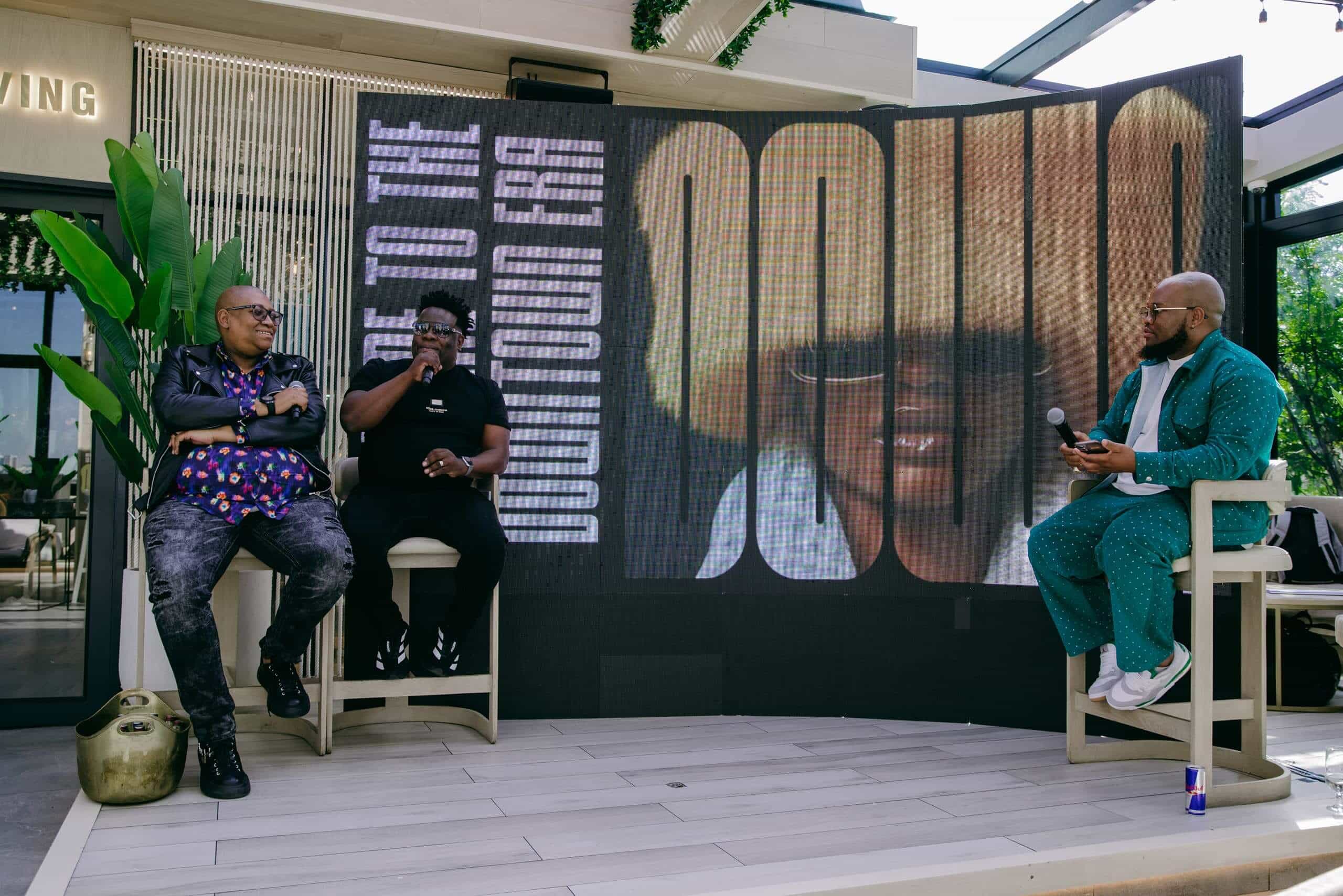 Three men sit and talk on a stage. Two are seated on the left, dressed in patterned shirts and dark pants, one holding a microphone. The third man, in a turquoise suit, sits on the right holding a microphone. Behind them is a large screen with stylized text and a woman's portrait.