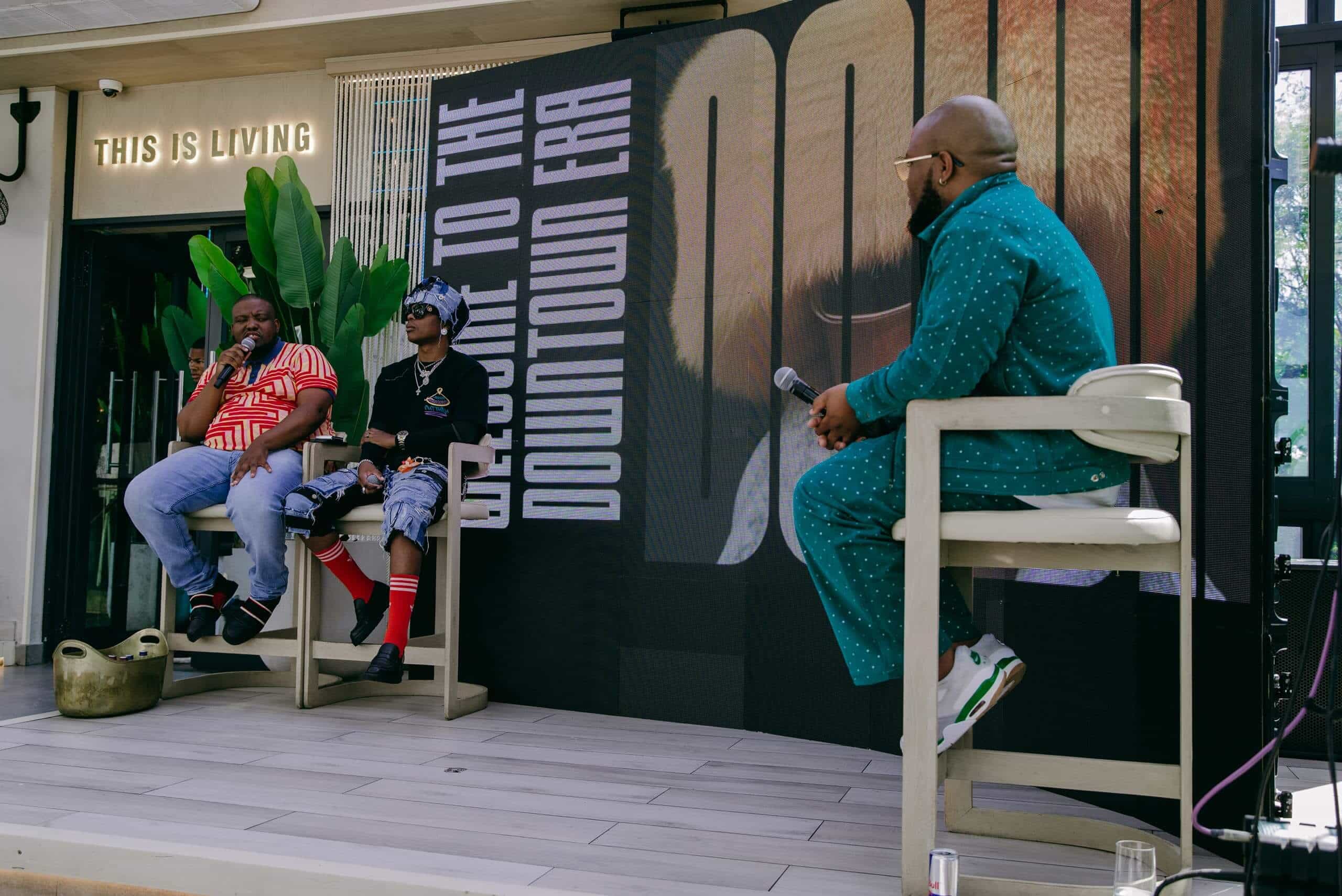 Three people are on a stage with &ldquo;Welcome to the Downtown Era&rdquo; on a large screen behind them. Two sit on the left, one in a red-striped shirt, the other in sunglasses and layered clothing. The third person, in a teal outfit, sits right holding a microphone.