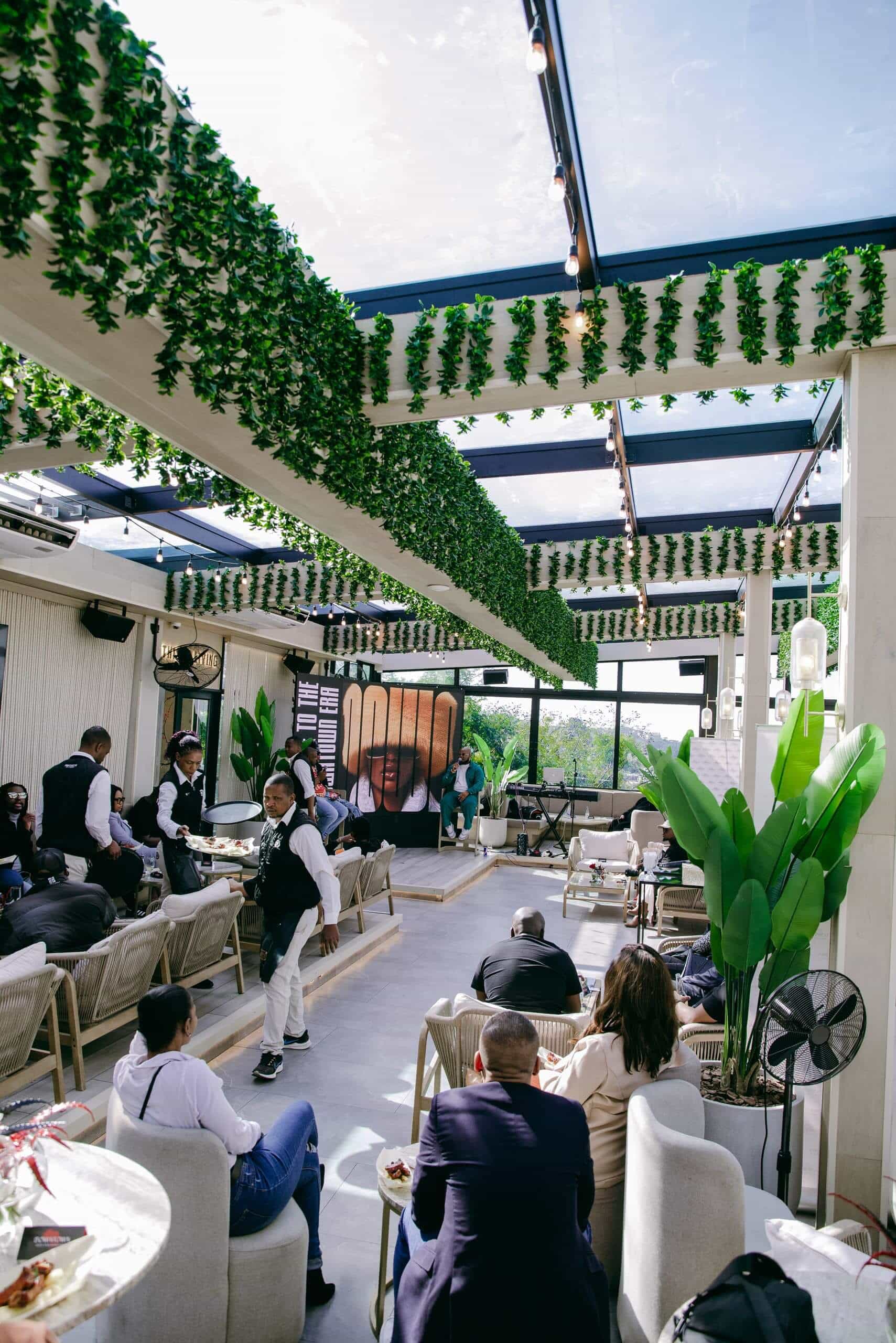 A modern rooftop lounge with hanging greenery, glass ceiling, and cushioned seating. People are seated, chatting, and eating while servers in black uniforms move around. Sunlight streams in, creating a bright, lively atmosphere. Large plants decorate the space.