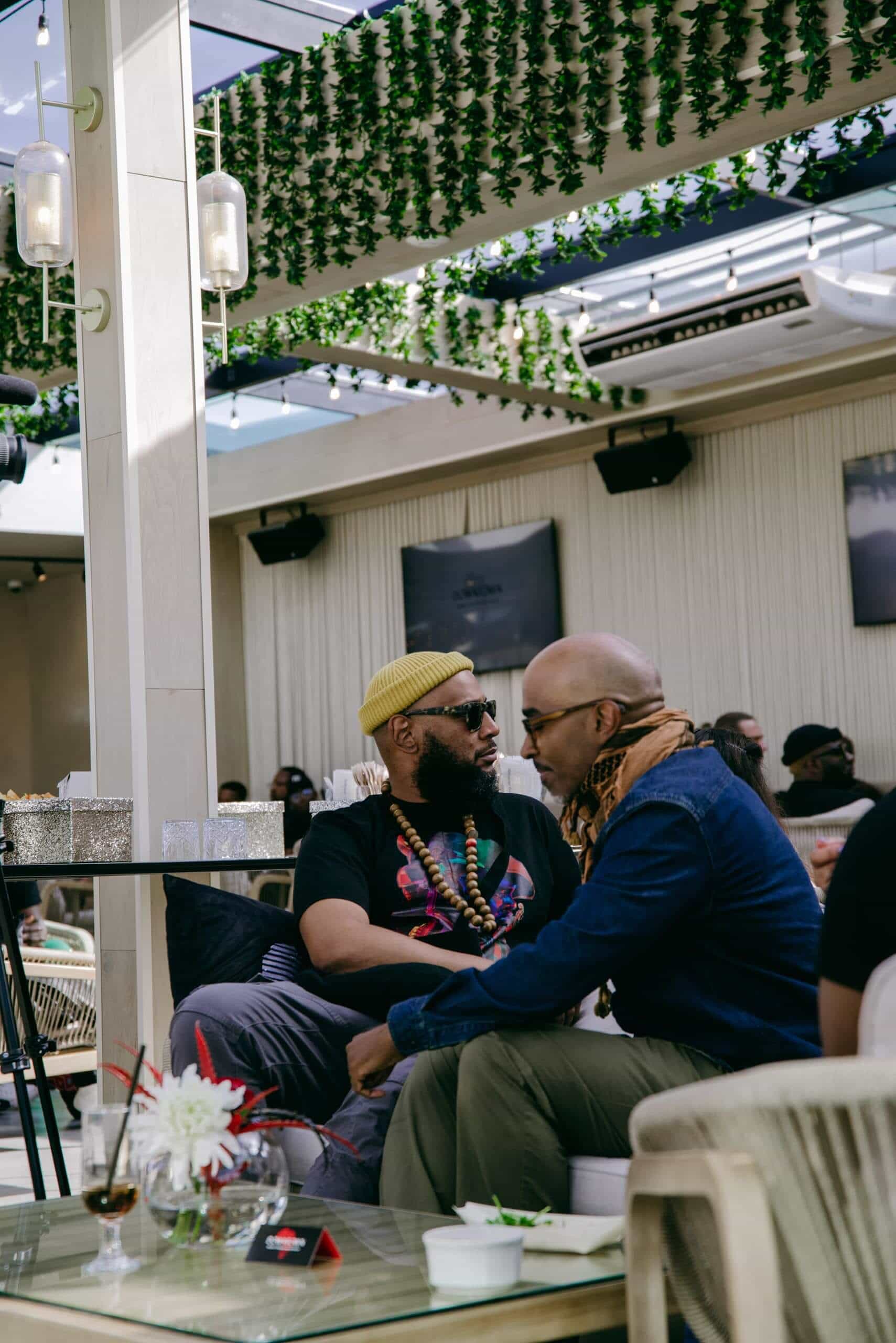 Two men sit closely together in conversation at a stylish caf&eacute; with greenery overhead. One wears a yellow knit hat, sunglasses, and colorful beads; the other wears glasses and a tan scarf. Both appear relaxed, surrounded by modern d&eacute;cor and other patrons.