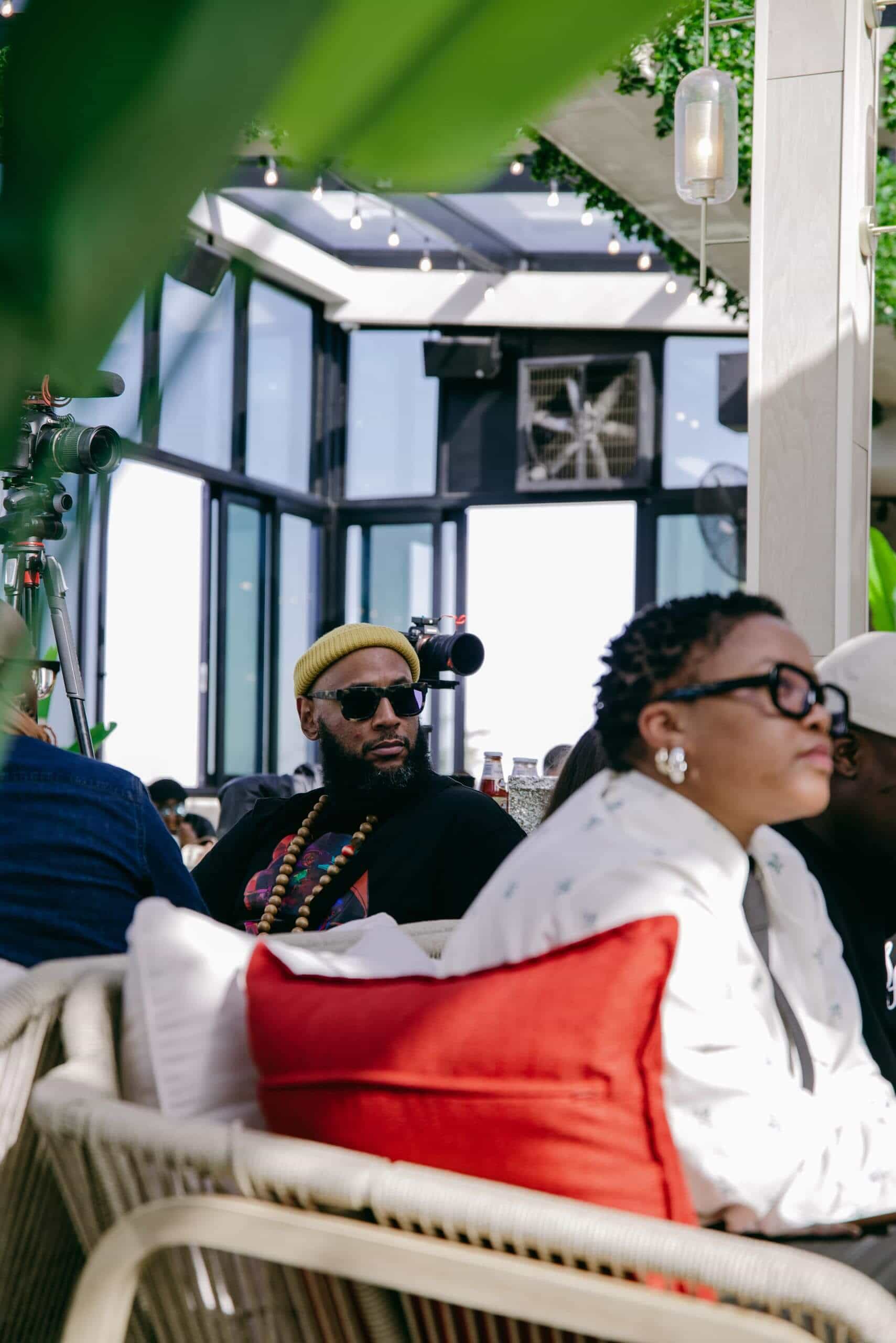 A man wearing a yellow beanie, sunglasses, and colorful beaded necklaces sits on a wicker chair with a red cushion, indoors near large windows and plants. Another person, out of focus, sits in the foreground. The setting is bright and modern.