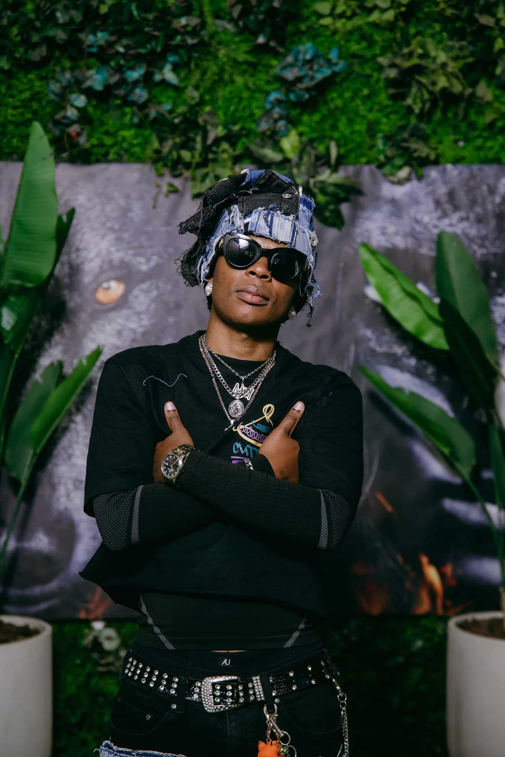 A person wearing sunglasses, layered necklaces, a black shirt, studded belt, and a denim headwrap stands with arms crossed in front of a leafy wall and artwork of a wolf's face. Two tall potted plants flank the subject.
