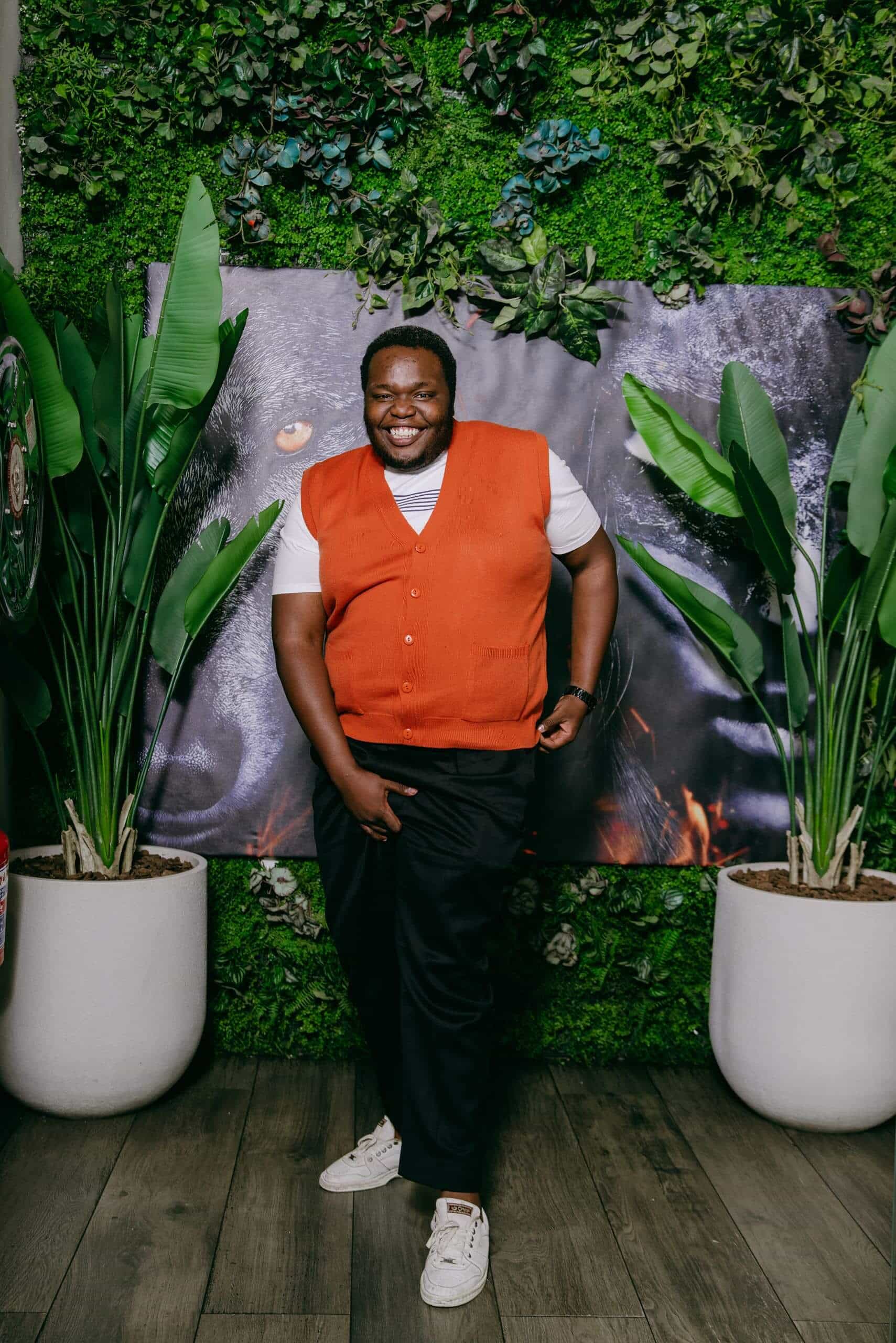 A smiling man in an orange vest and white t-shirt stands in front of a green plant wall and a black poster. He wears dark pants and white sneakers, with potted plants on either side of him, on a wood floor. He stands relaxed, pointing one finger down.