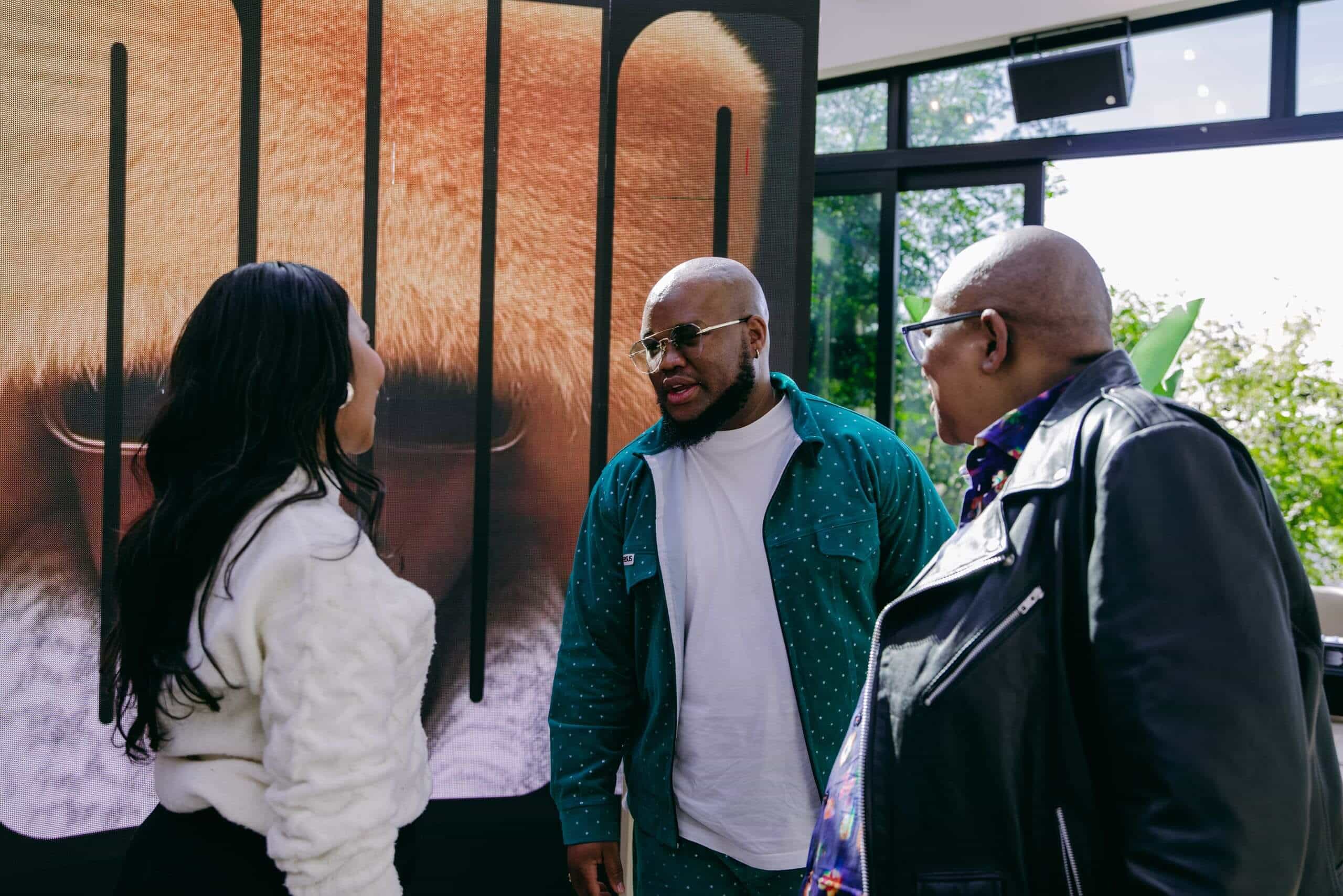 Three people stand indoors having a conversation. One woman in a white sweater faces two men, one in a green outfit and another in a black leather jacket. Behind them is a large poster with an animal&rsquo;s fur and nose and bold vertical letters.