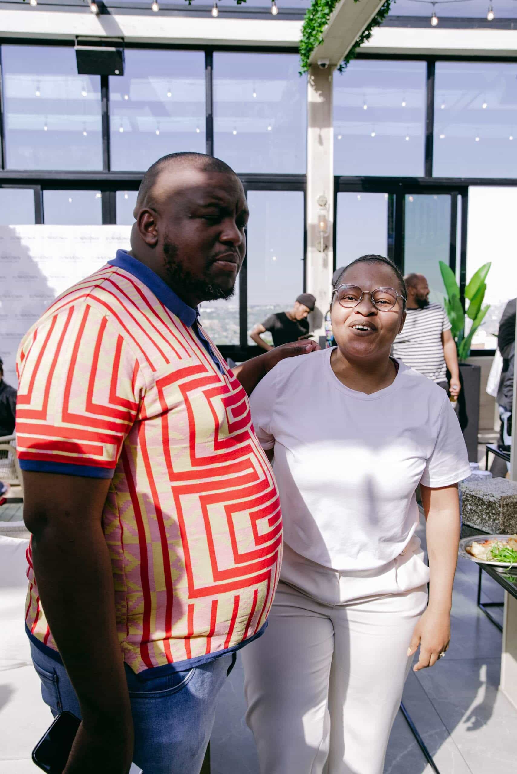 Two people stand indoors smiling. The man on the left wears a colorful red and yellow geometric-patterned shirt and jeans. The woman on the right wears glasses, a white t-shirt, and beige pants. Sunlight streams in from glass windows behind them.