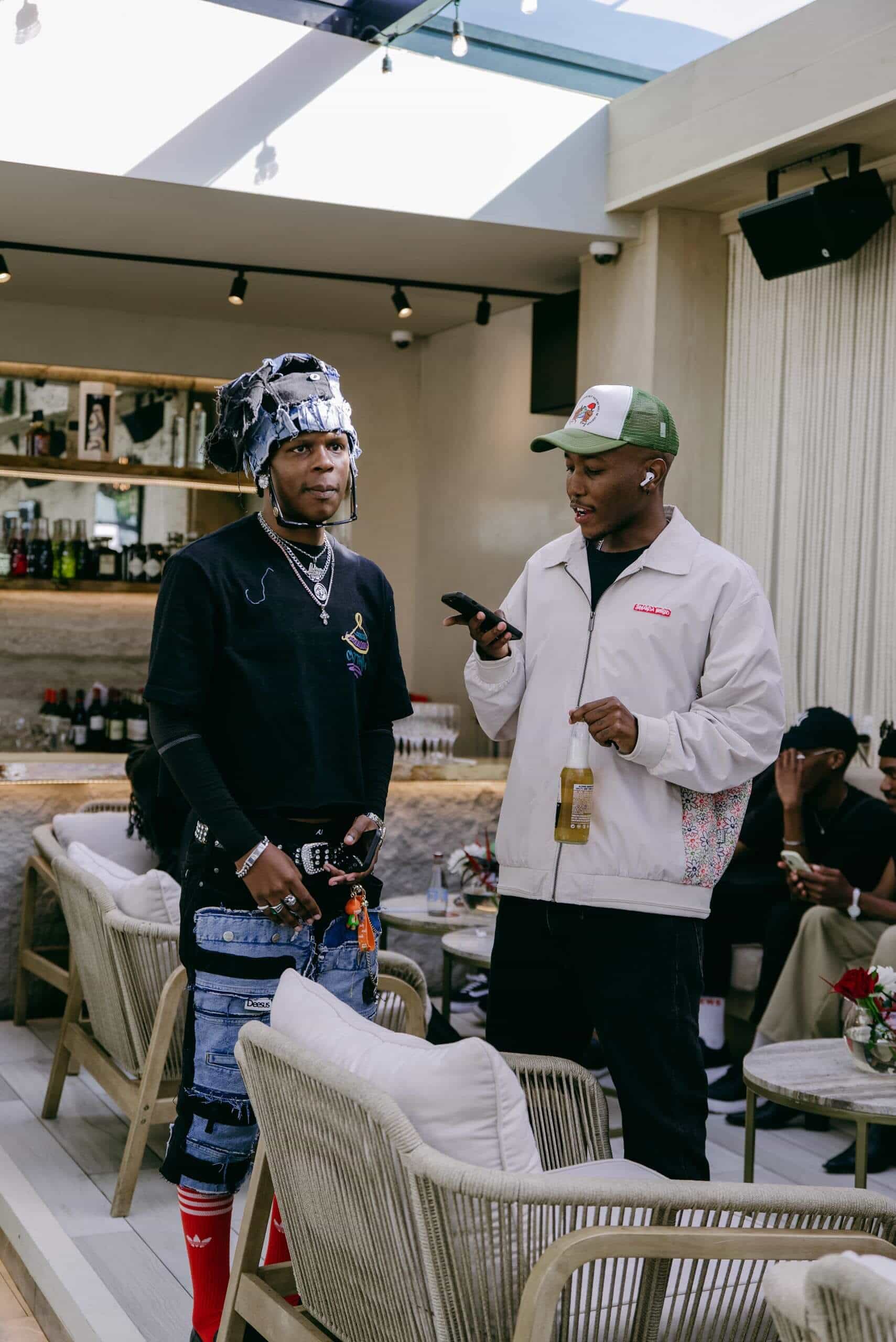 Two men stand indoors at a stylish lounge. One wears layered silver chains, a helmet-like headpiece, graphic sweatshirt, patched jeans, and red socks. The other wears a cap, light jacket, holds a beer, and uses a phone. Bar and seated people are visible in the background.