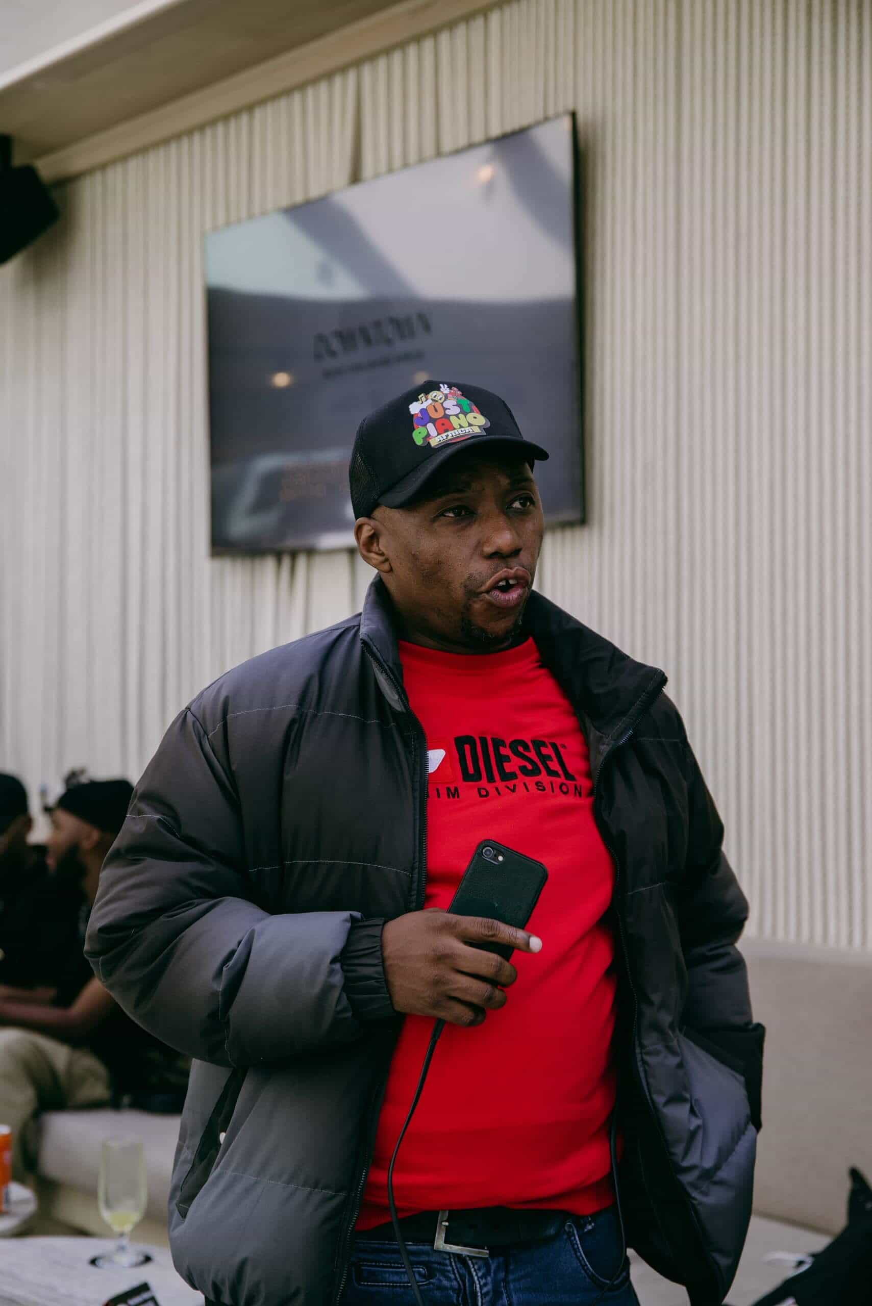 A man wearing a black cap, gray puffer jacket, and red Diesel shirt holds a smartphone and speaks. He stands indoors near a beige couch, with a wall-mounted TV and other people blurred in the background. A drink sits on a table nearby.