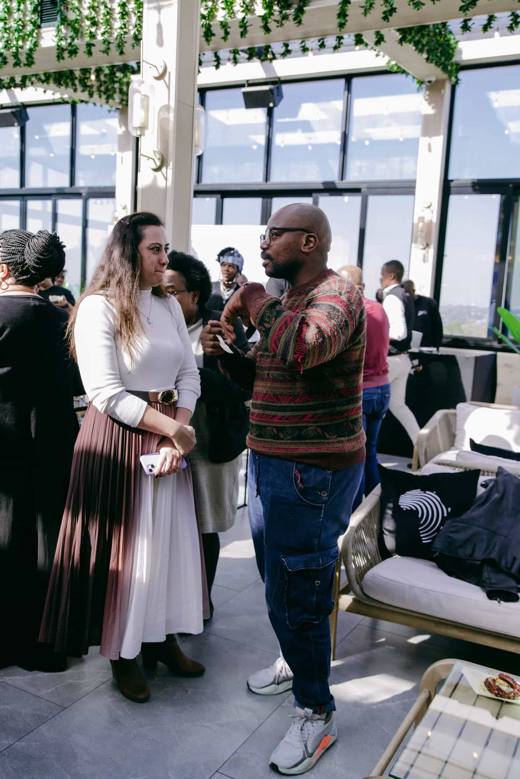 A woman in a long-sleeved white top and pleated brown skirt stands talking with a man in glasses, a patterned sweater, and blue cargo pants at an indoor event. Other people mingle in the background amid modern decor and large windows.