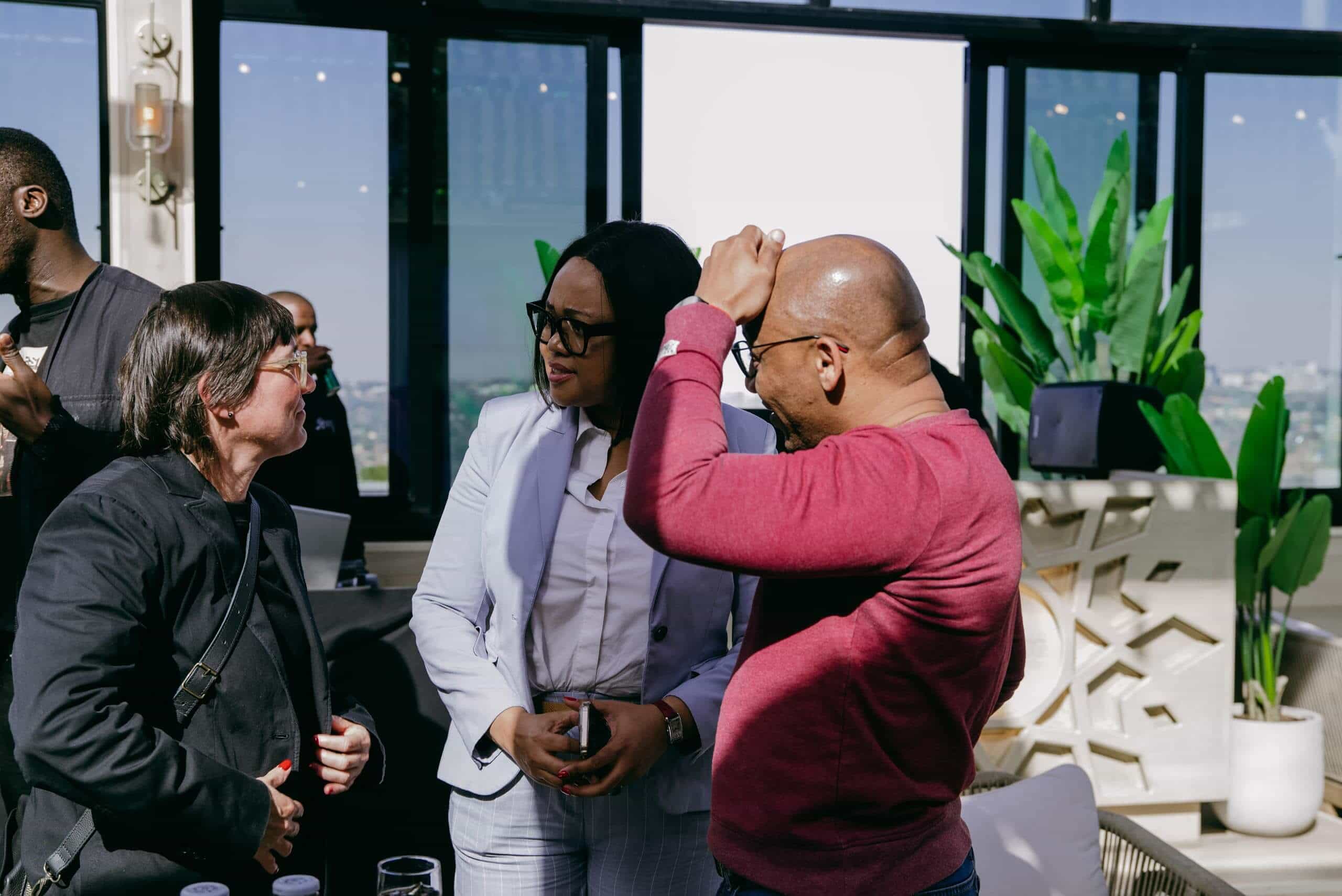 Three adults are chatting indoors near large windows with city views. One woman wears a white suit, another wears glasses and a black jacket, and a man in a red shirt gestures with his hand. A bright plant and sunlit decor are in the background.