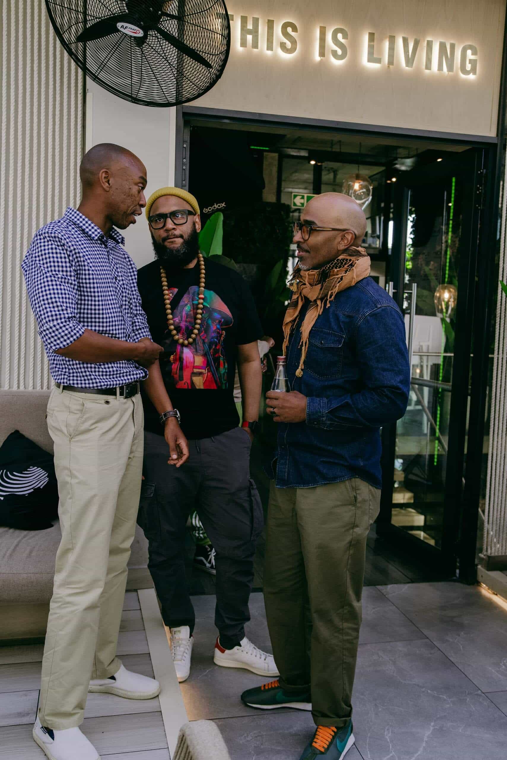Three men stand and chat in front of a modern building entrance with a glowing &ldquo;THIS IS LIVING&rdquo; sign above the door. One man wears a checkered shirt and khakis, another has a yellow beanie and graphic tee, and the third sports glasses, a scarf, and a blue shirt.