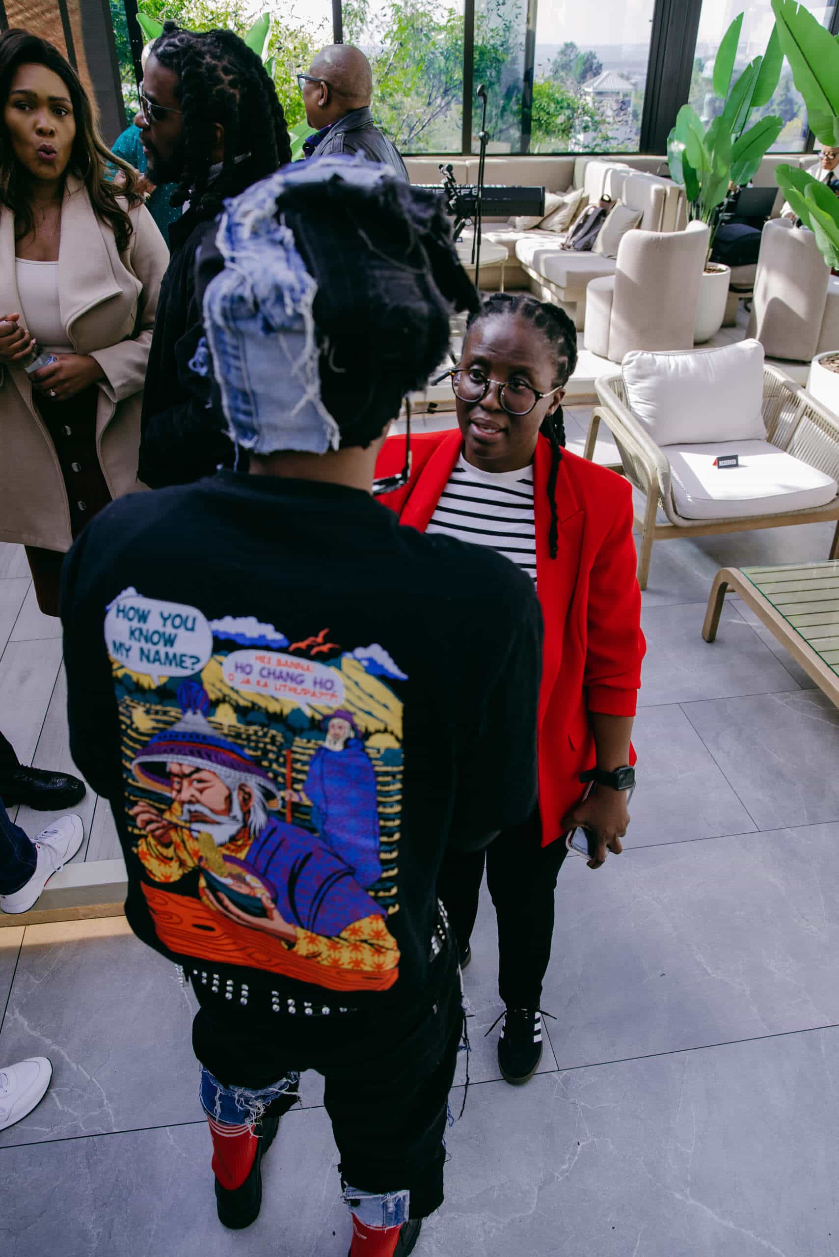 A person in a red blazer and glasses stands indoors, facing another person wearing a graphic hoodie and denim head wrap. Other people are nearby, with plants and modern furniture in the background. The atmosphere appears casual and social.
