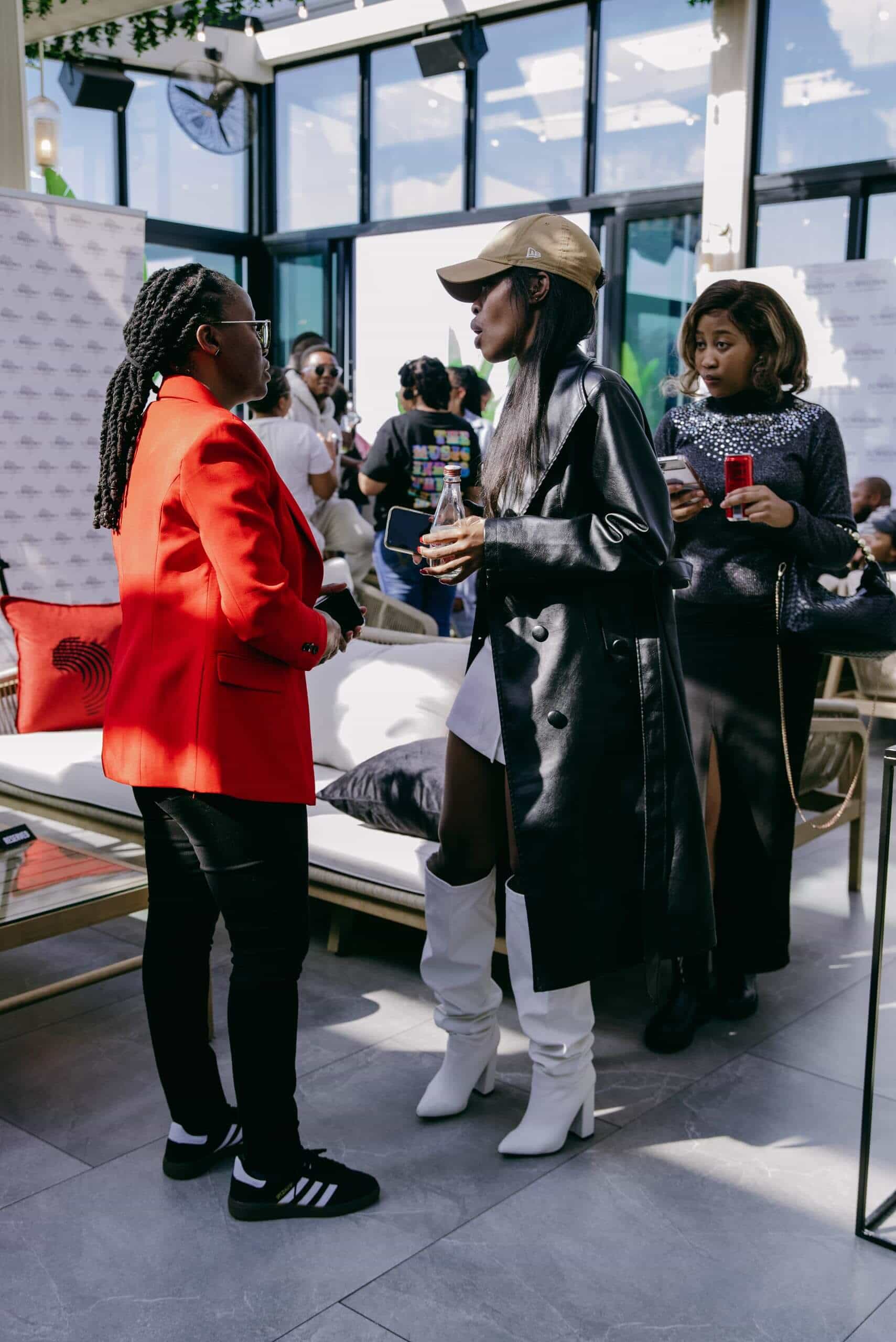 Two women stand facing each other indoors. One wears a red blazer, black pants, and Adidas sneakers. The other wears a black leather coat, white boots, a gold cap, and holds a drink. A third woman in black stands behind them, holding a phone and a red cup.