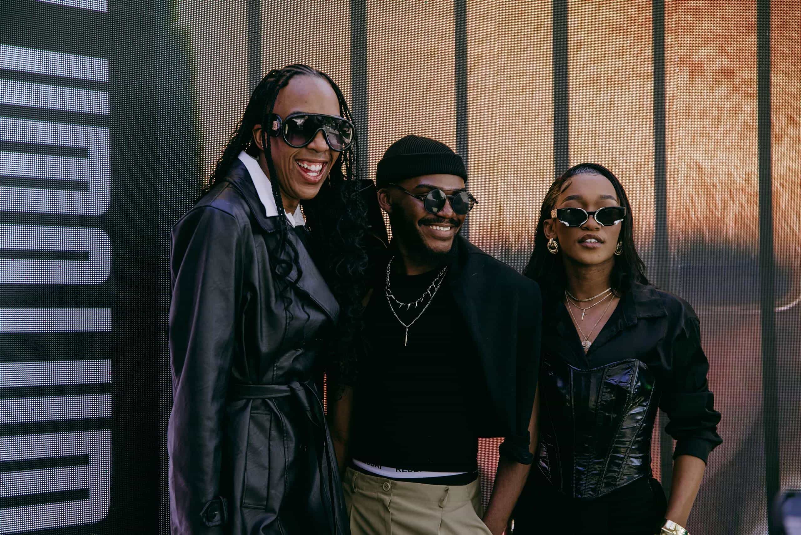 Three stylish people pose and smile together wearing fashionable black outfits and sunglasses. They stand in front of a modern backdrop with vertical dark lines and part of a large, blurred image. The mood is upbeat and confident.