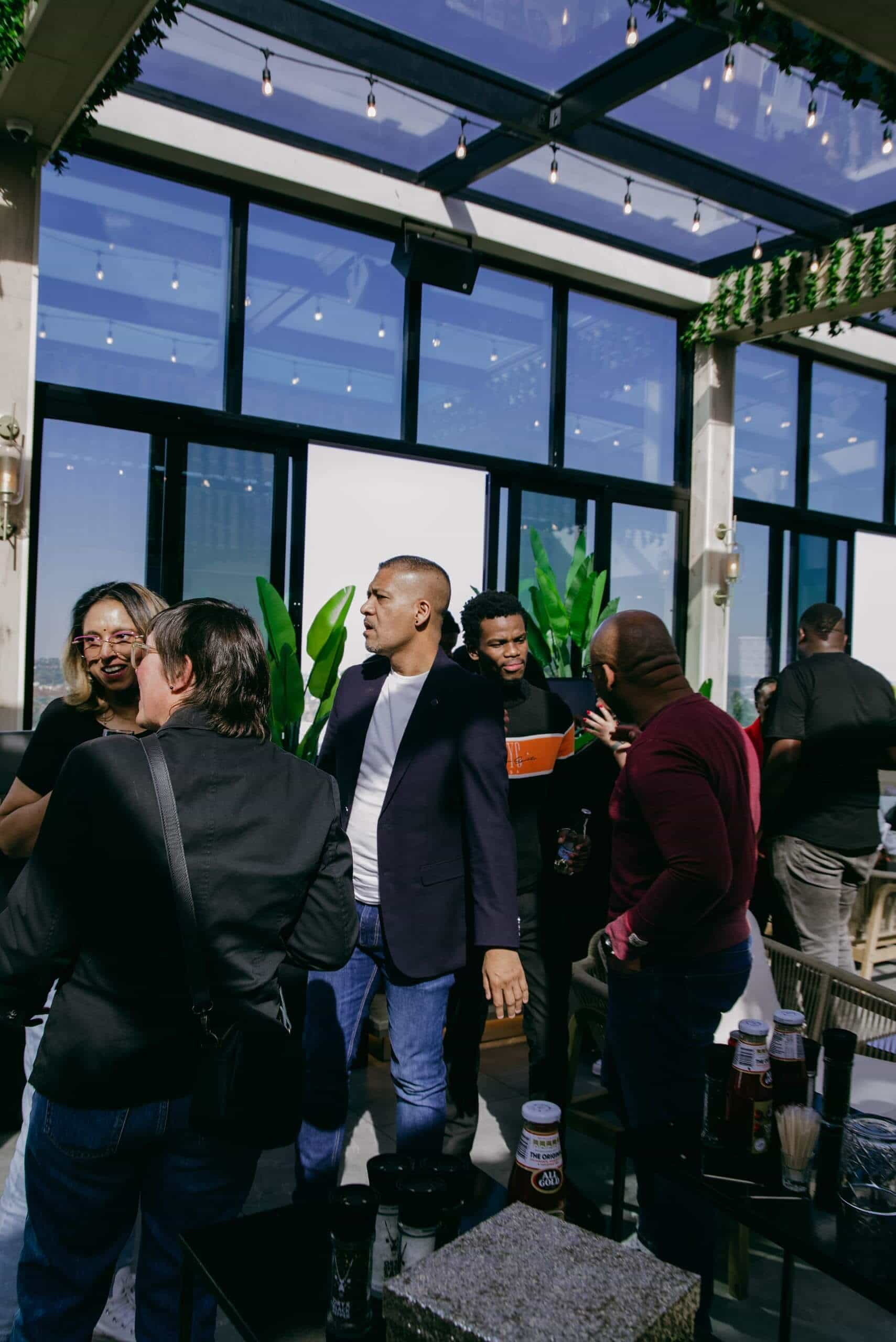 A group of people socialize indoors by large windows with sunlight streaming in. Some are smiling and talking, one holds an orange box. Plants and string lights decorate the modern space. Bottles and drinks sit on a nearby table.