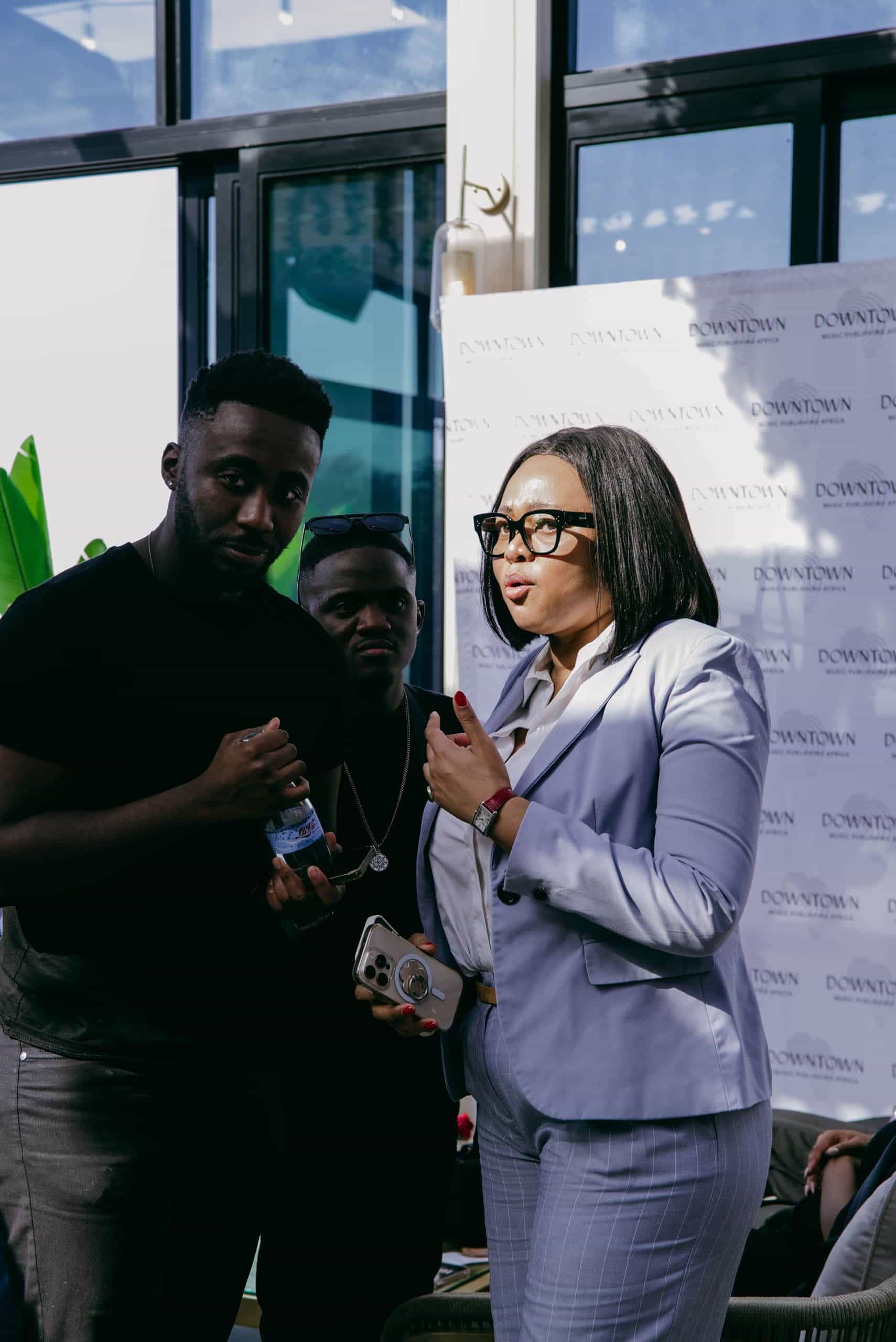 Three people stand together indoors. A woman in a light blue suit and glasses holds a phone and gestures while talking. Two men, one in a black shirt and another wearing sunglasses, stand nearby. There is a 