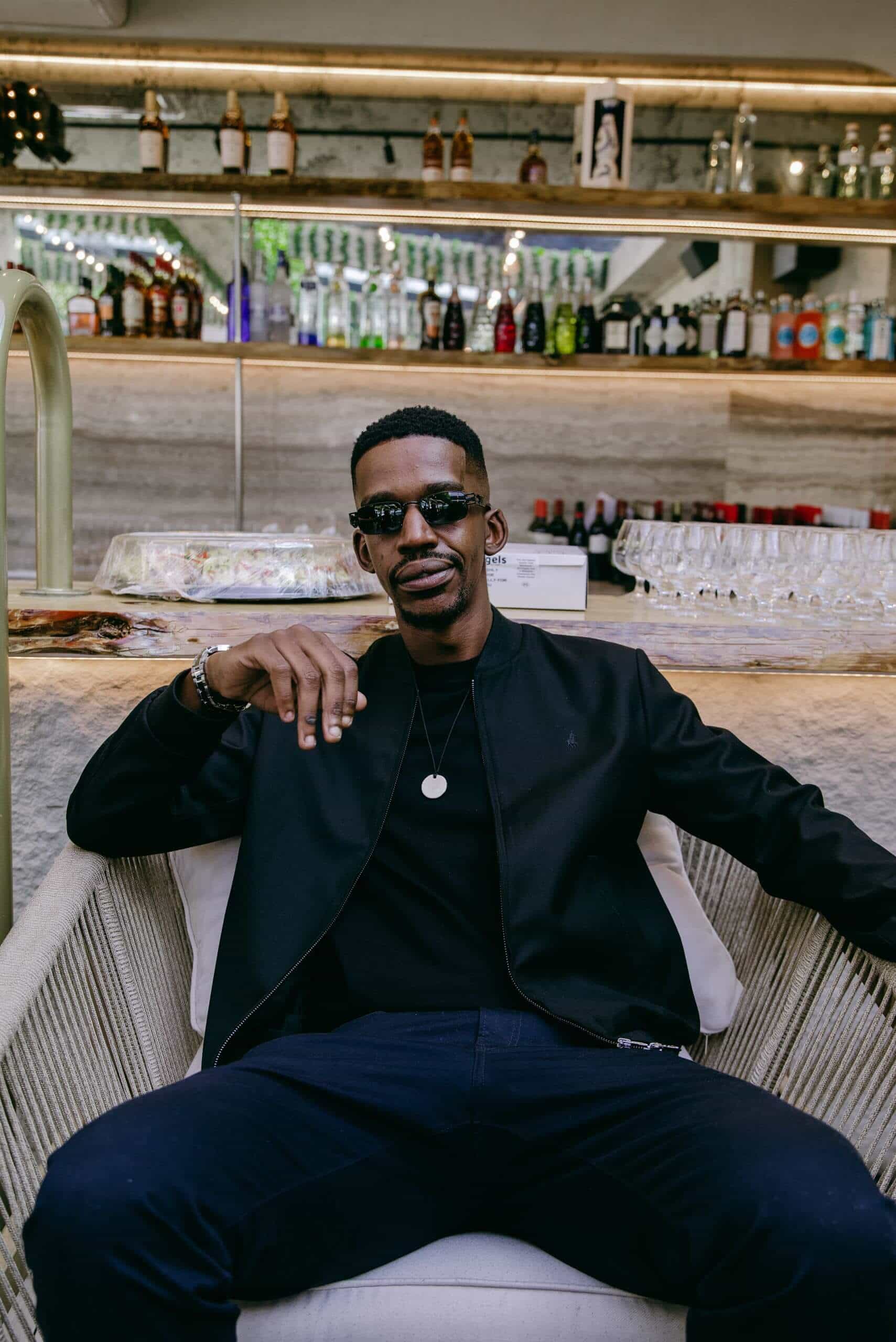 A man with short hair and sunglasses sits confidently in a woven chair, wearing a black jacket, black shirt, dark pants, and a necklace. Behind him is a bar with shelves full of assorted bottles and glassware. The setting appears modern and stylish.