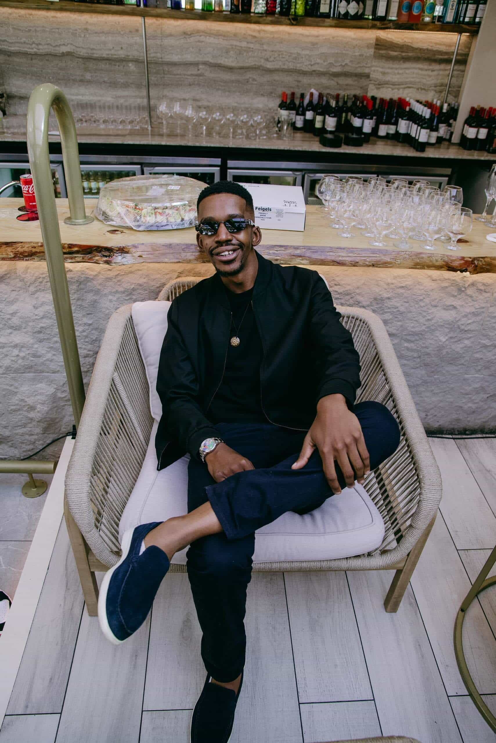 A man wearing sunglasses, a black jacket, pants, and blue loafers sits cross-legged on a cushioned chair, smiling at the camera. Behind him is a bar counter with glasses, wine bottles, and snacks arranged neatly.