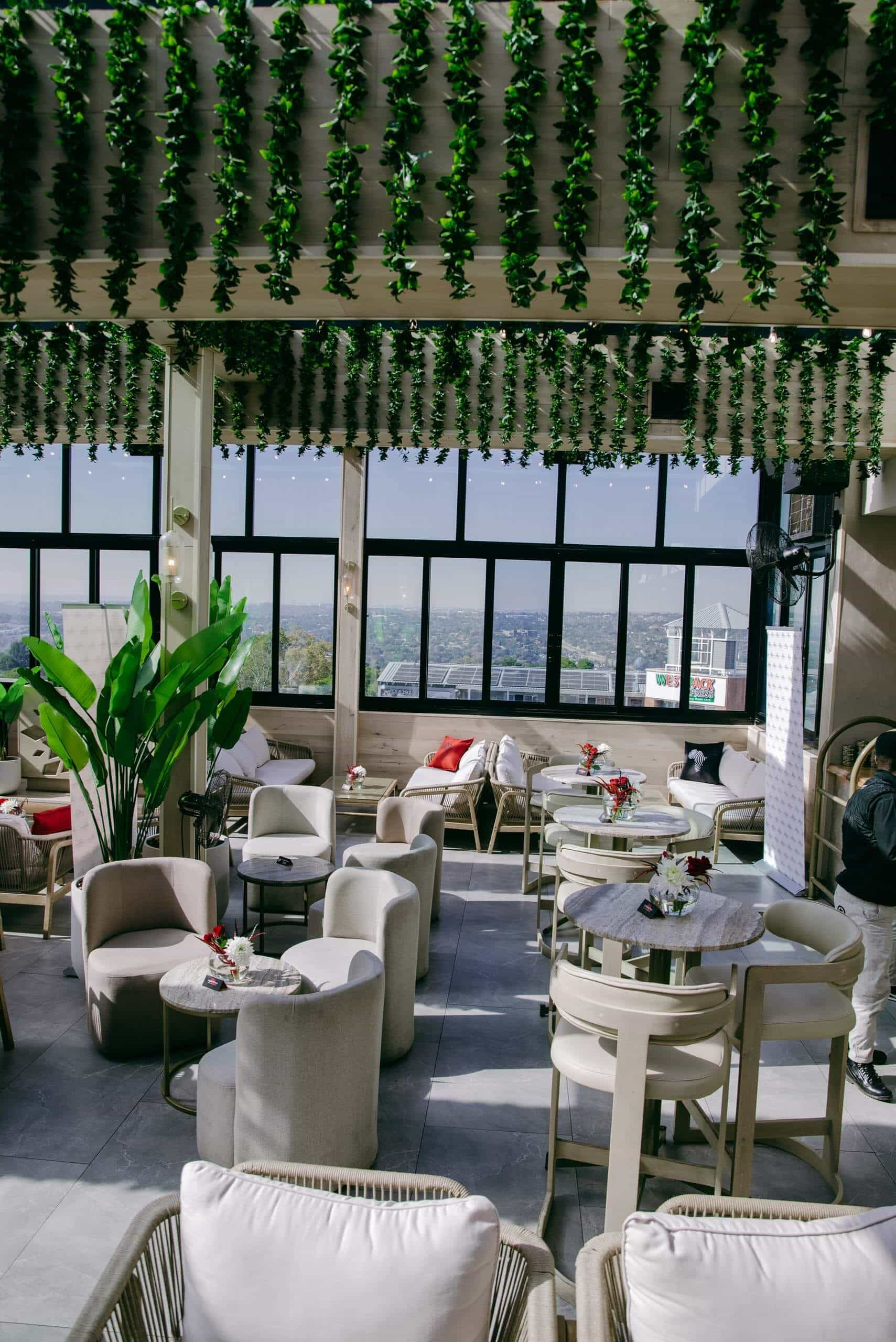 A bright, modern caf&eacute; with white cushioned chairs and round marble tables. Green vine decorations hang from the ceiling, and large windows let in sunlight, offering a scenic view outside. Potted plants and red accent pillows complete the fresh, inviting space.