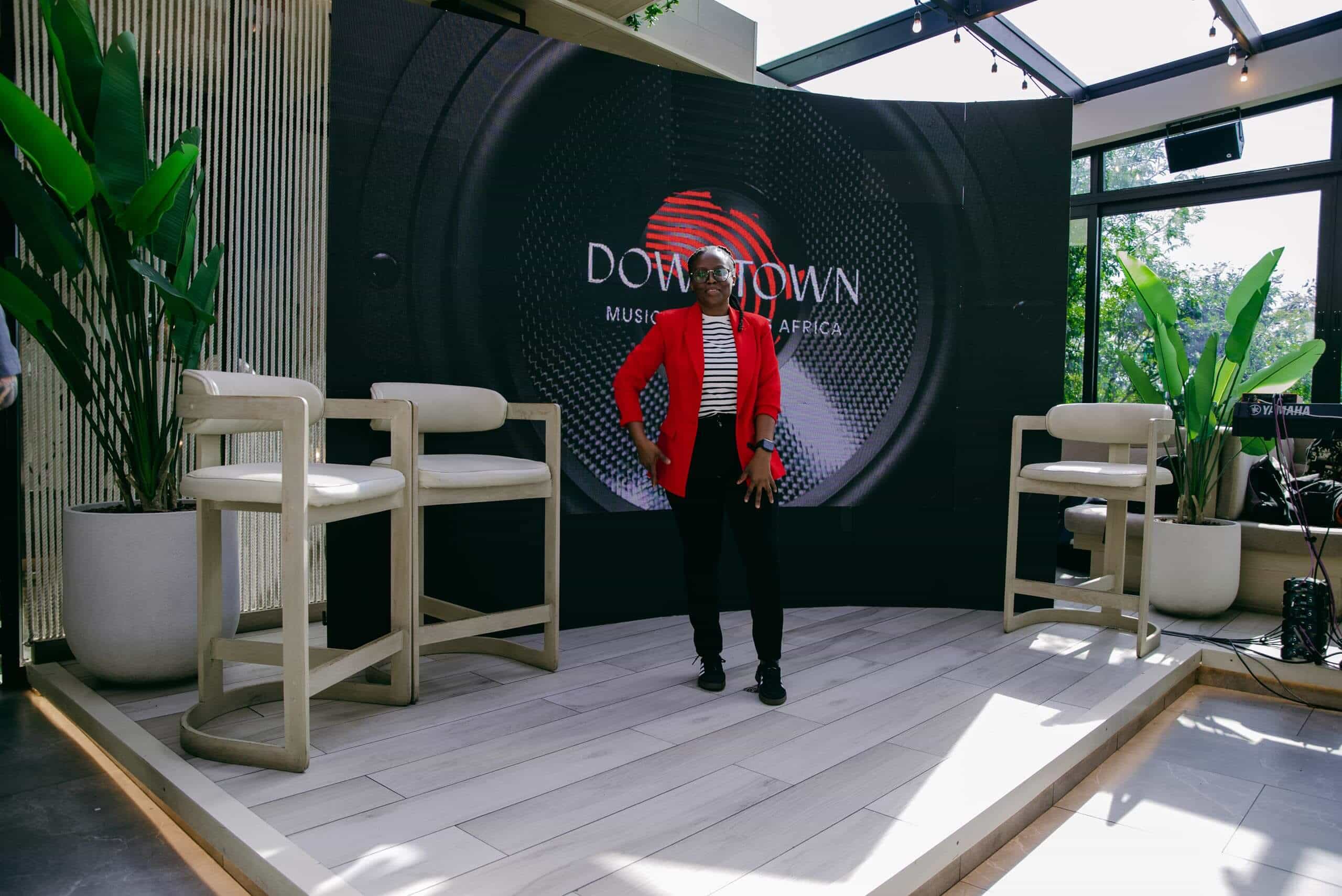A person wearing a red blazer, black pants, and a striped shirt stands on a small stage between two white chairs. Behind them is a large screen displaying the words 