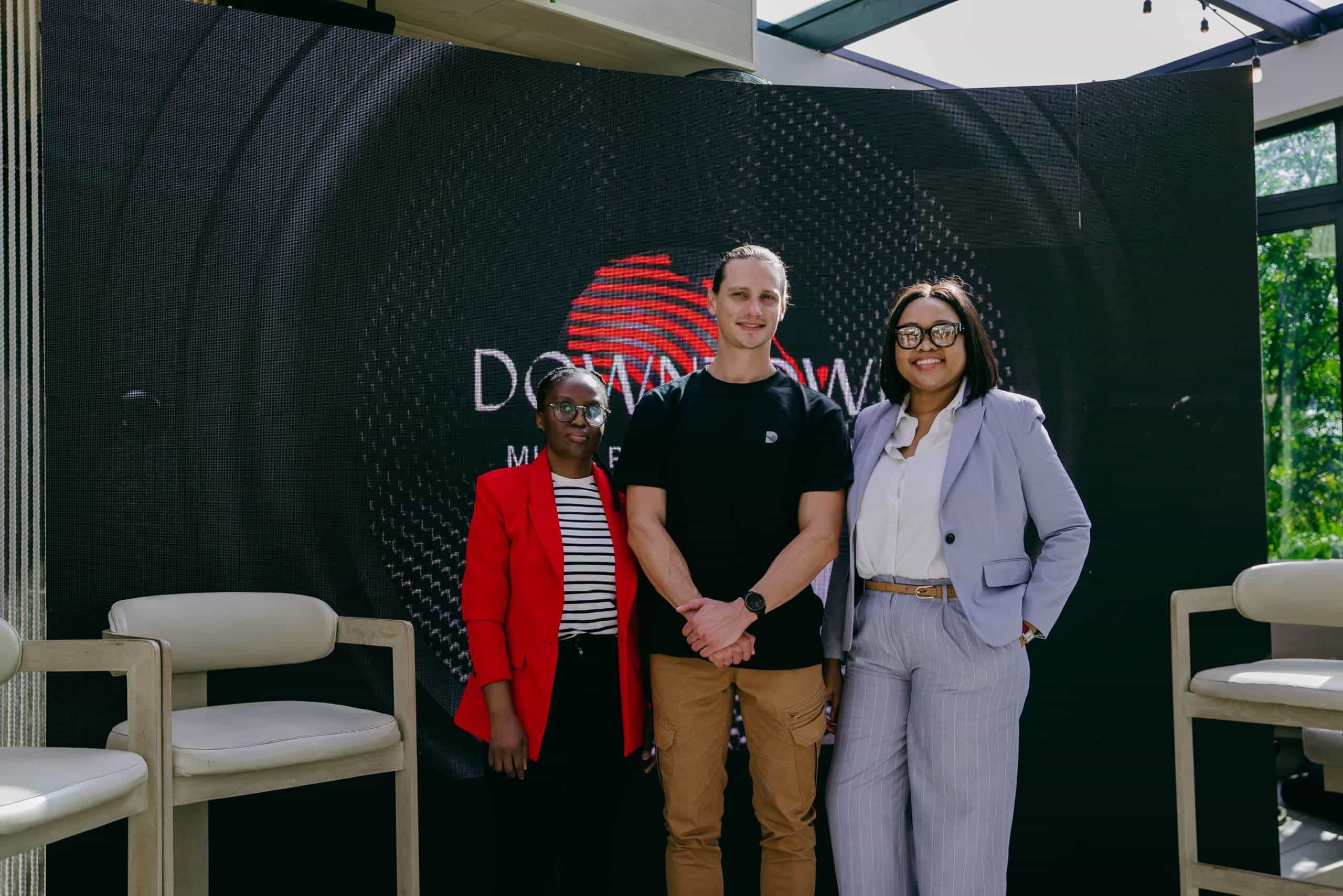 Three people stand in front of a large black display screen with a red logo and the words &ldquo;Downtown Masterclass.&rdquo; The person on the left wears a red blazer, the person in the middle is in a black t-shirt, and the person on the right wears a light suit and glasses.