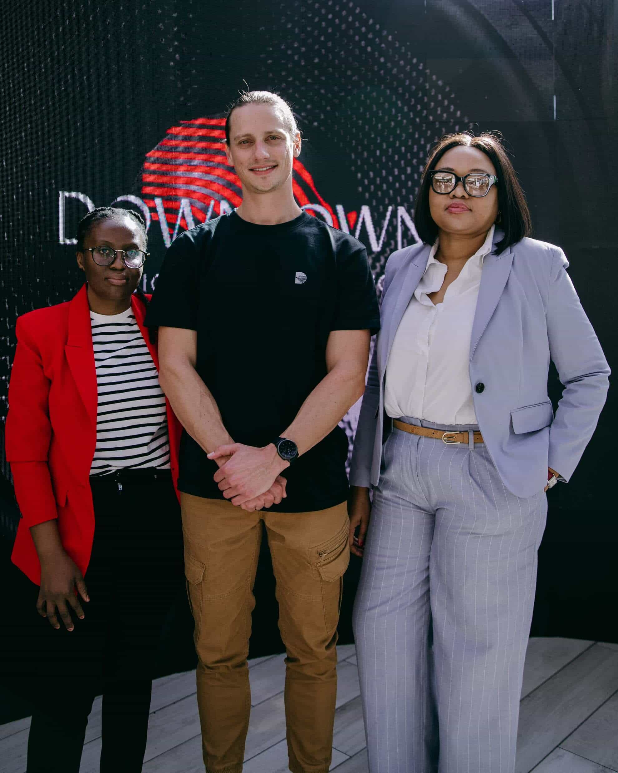Three people stand side by side, posing for a photo. The person on the left wears glasses, a red blazer, and striped shirt. The person in the middle wears a black t-shirt and brown pants. The person on the right wears glasses, a lilac suit, and a white blouse.