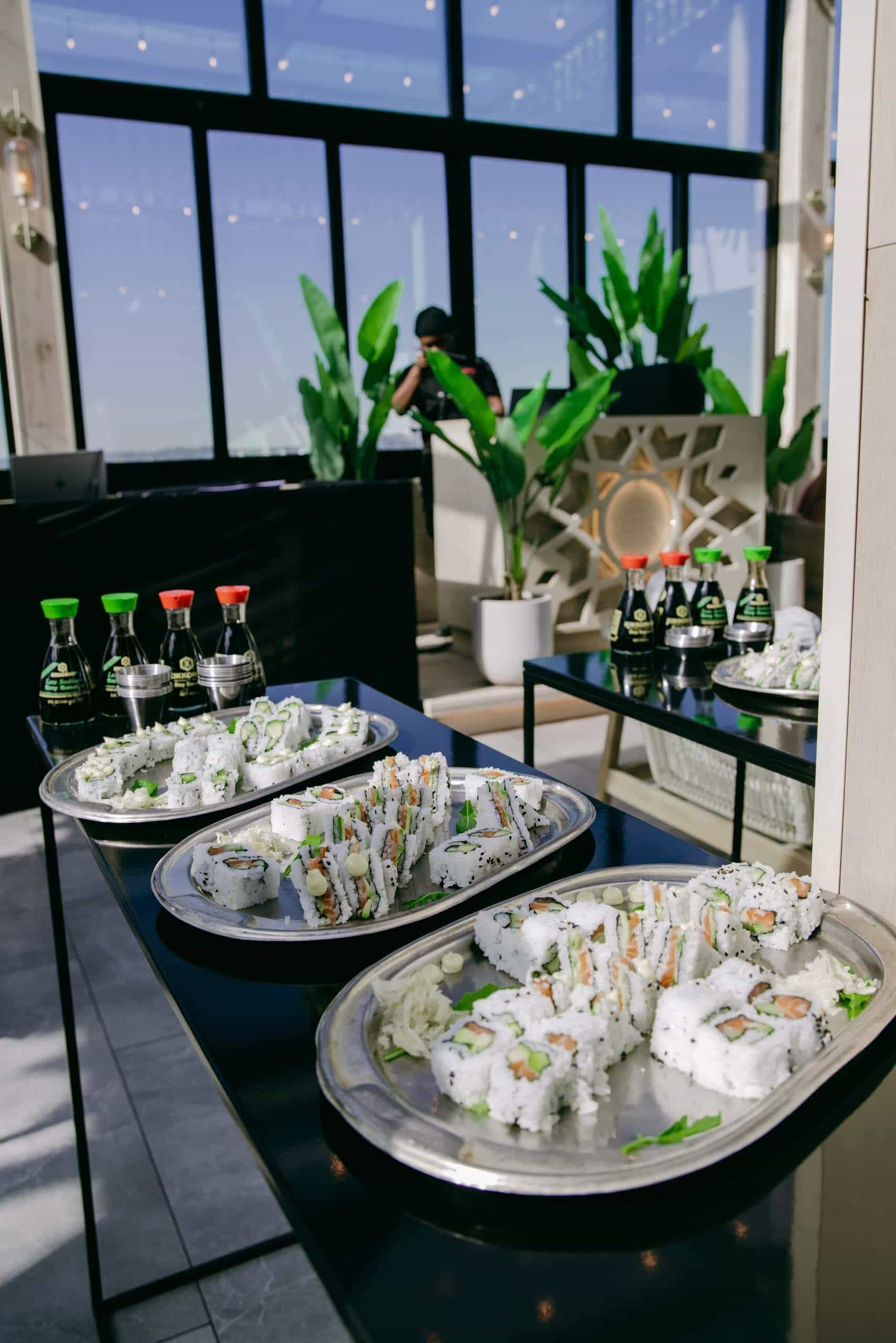 Three silver platters of assorted sushi rolls are displayed on a black table, garnished with wasabi and ginger. In the background, bottles of soy sauce are neatly arranged, with large green plants and a modern interior with tall windows visible.