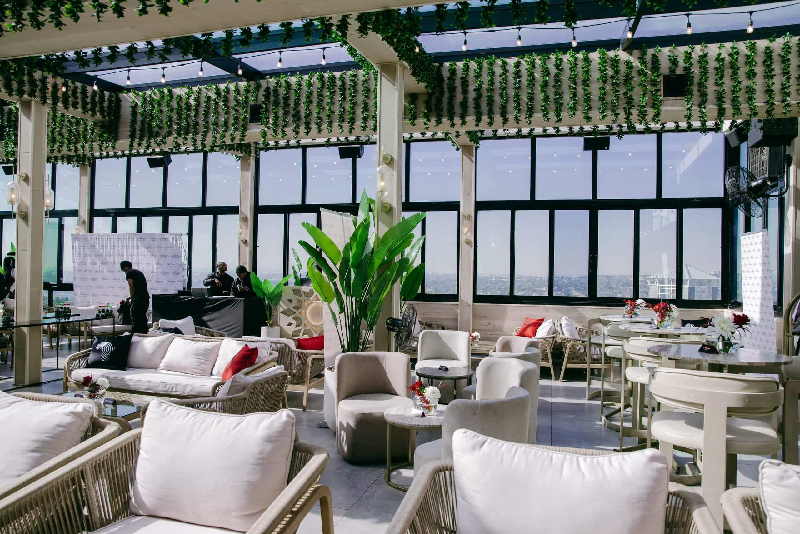 A bright, modern rooftop lounge with large windows, white cushioned chairs, small tables, green plants, and hanging greenery. Red and white pillows accent the seating. Three people set up a table in the background near a step-and-repeat banner.