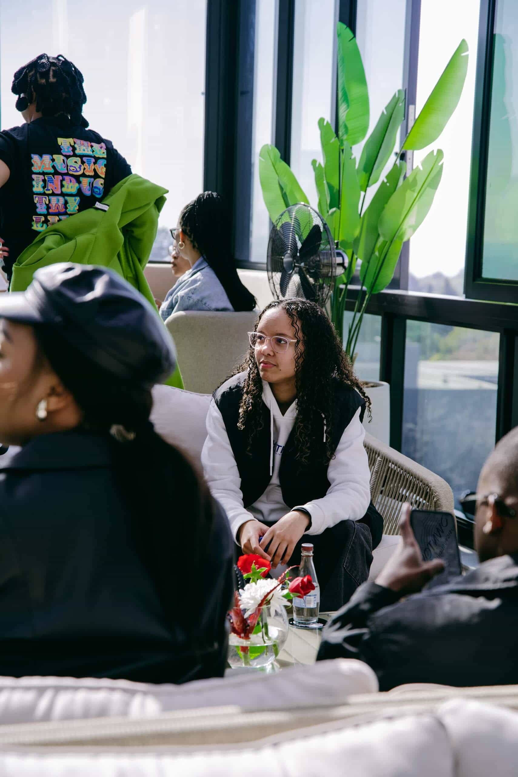 A young woman with glasses and long curly hair sits on a light-colored woven chair indoors, surrounded by other people. She wears a black and white jacket. There are plants, a table with flowers and a soda bottle, and large windows letting in natural light.