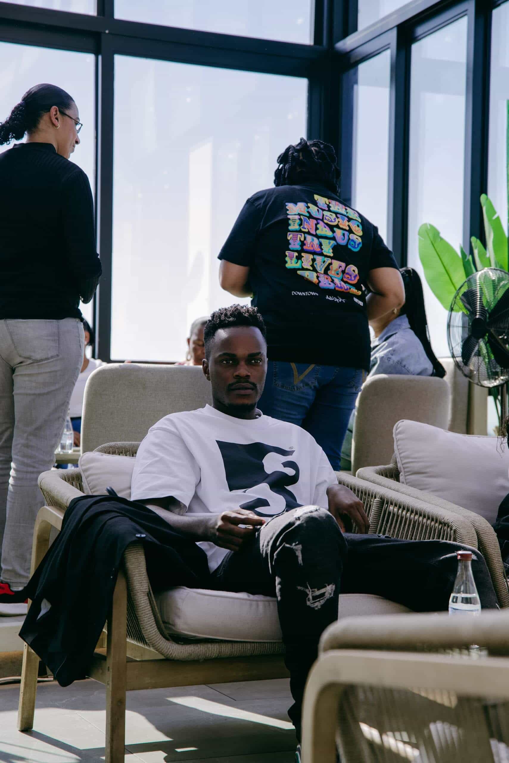 A man sits on a beige armchair inside a sunlit room with large windows. He wears a white and black graphic t-shirt and holds a phone. Two women stand nearby, one in a black shirt facing away. Potted plants and other people are visible in the background.