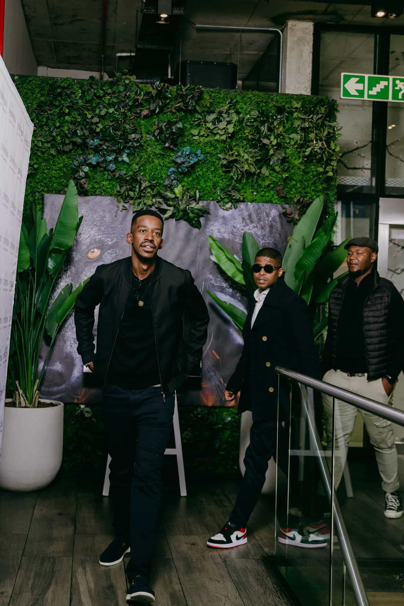 Three stylishly dressed men walk indoors along a wooden floor beside a glass railing. Behind them is a lush green wall with large plants and a partial image of an animal&rsquo;s eye. Emergency exit signs are visible above. The atmosphere is modern and trendy.