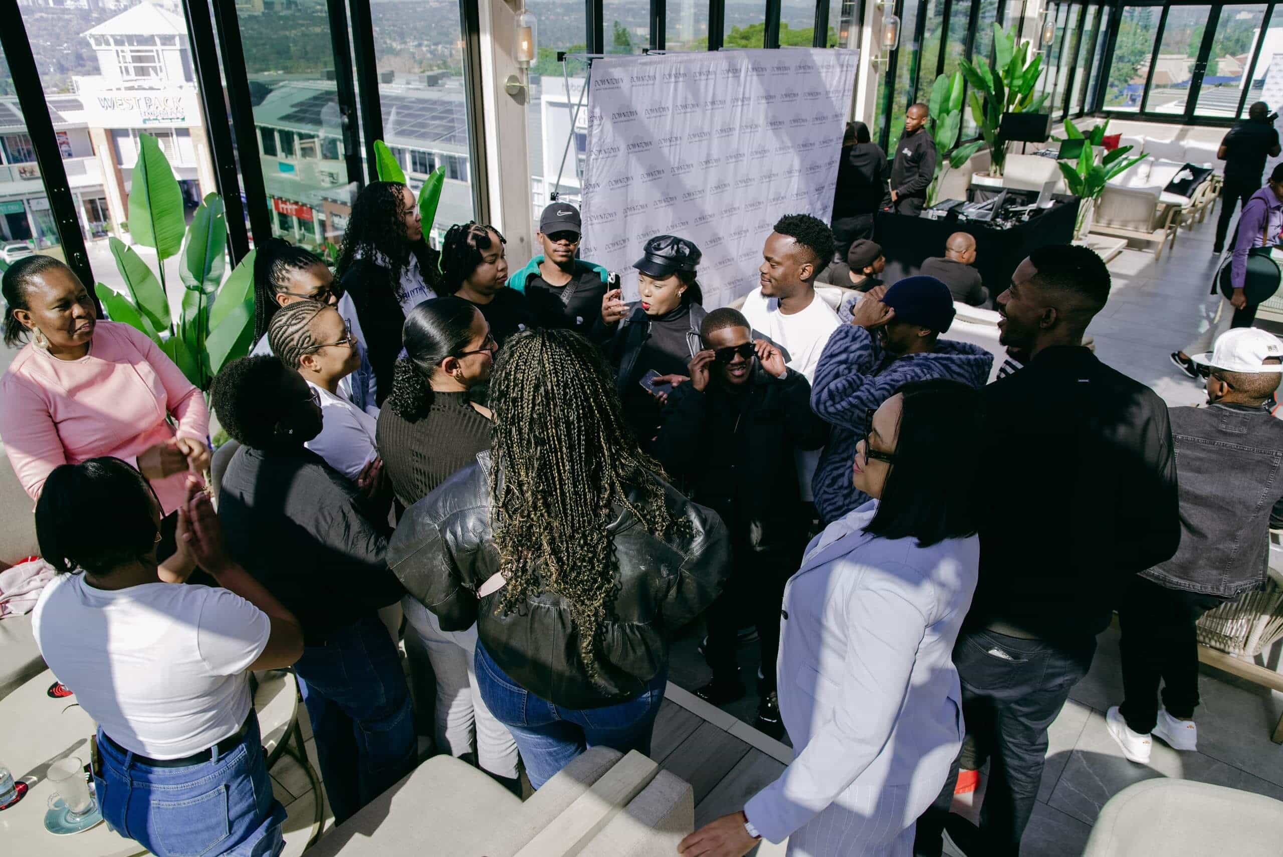 A diverse group of people, mostly young adults, gather indoors around a man speaking. Some listen attentively, others chat or use phones. Large windows show a city view outside. Potted plants and a step-and-repeat banner are in the background, suggesting an event or meetup.