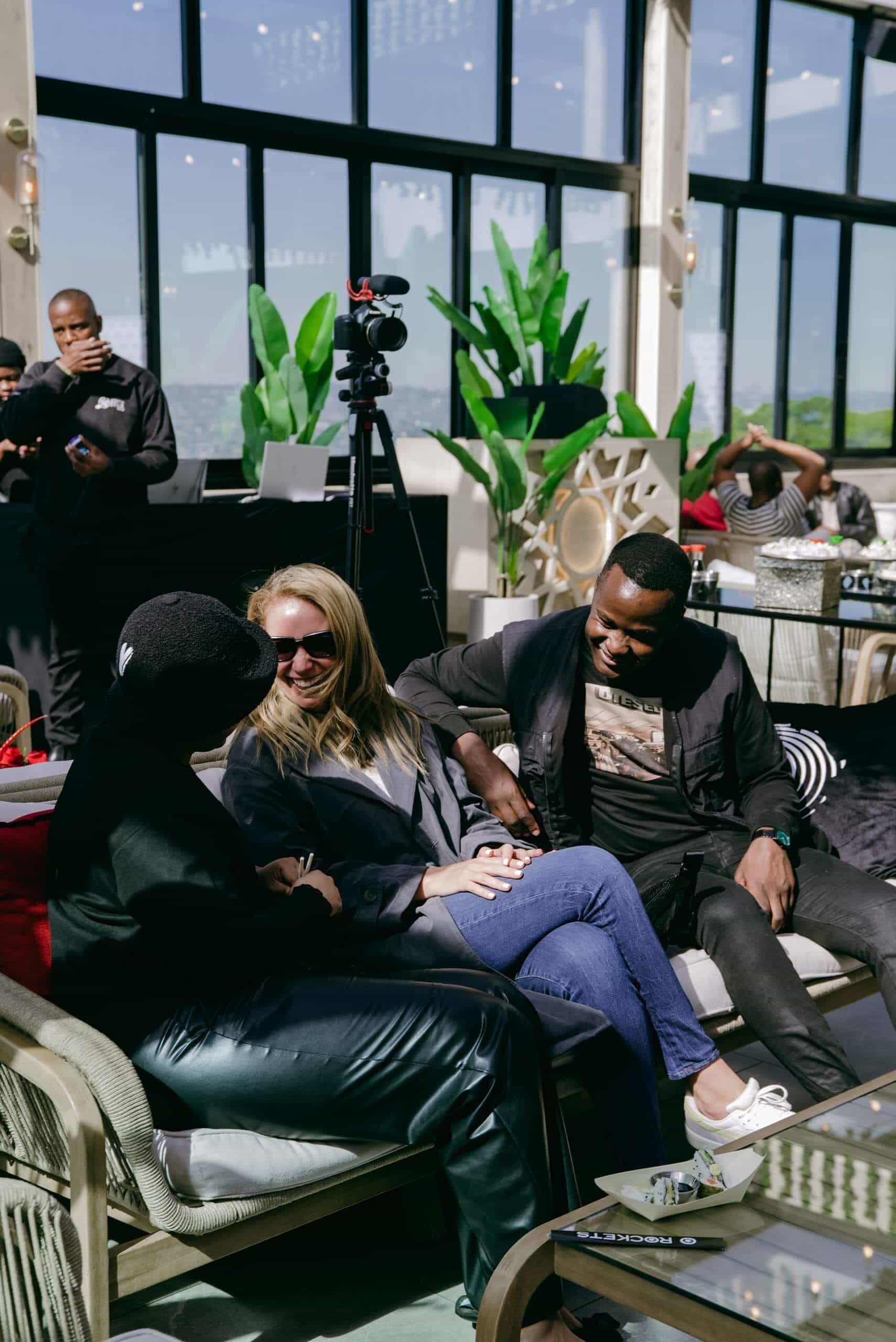 Three people sit on a sofa indoors, chatting and smiling. A woman in sunglasses sits between a man and a woman in black. In the background, a man uses a phone and another stands near a camera tripod. Large windows and green plants decorate the bright space.