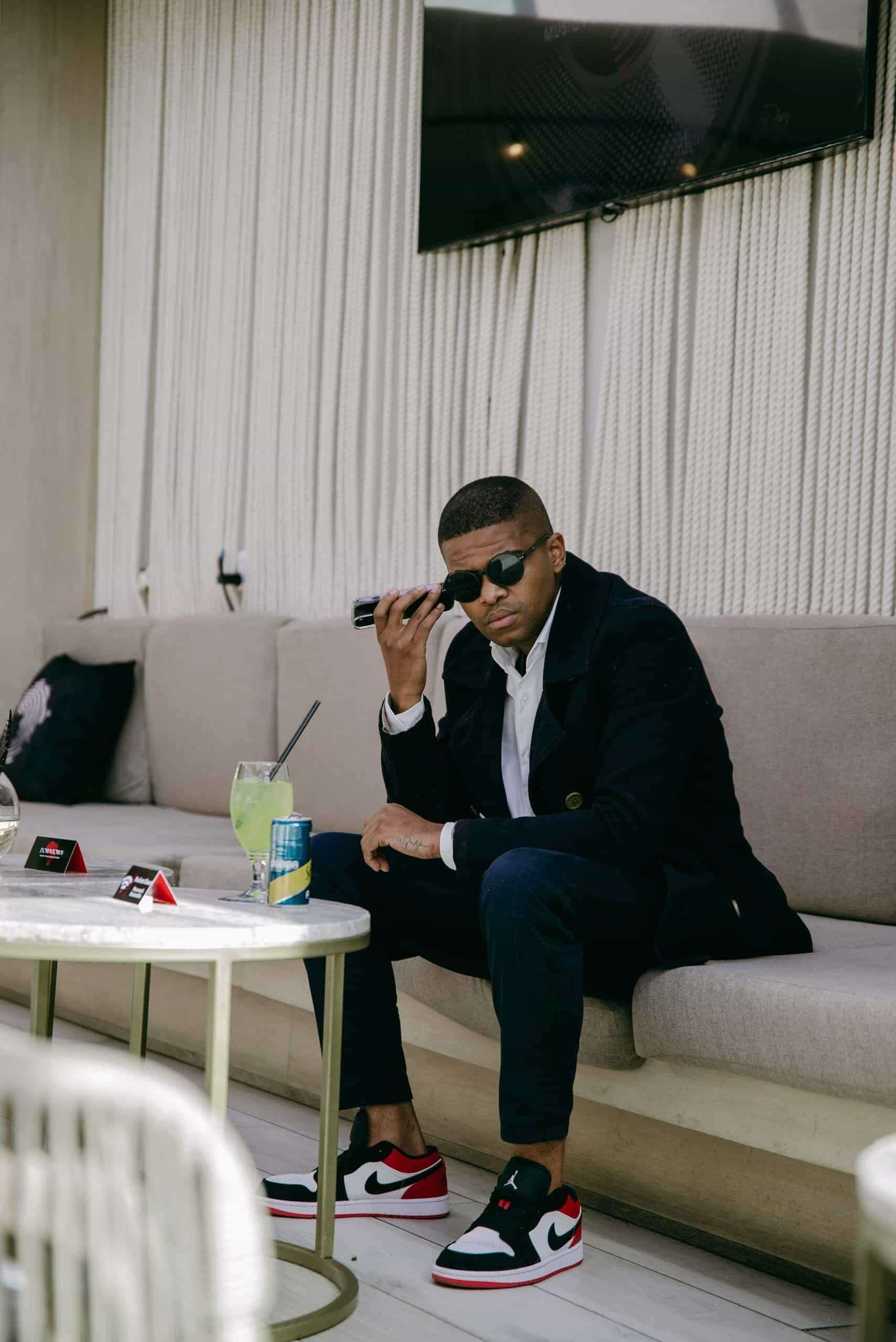 A man in a dark coat, white shirt, and sunglasses sits on a beige outdoor sofa. He holds a phone to his ear. He wears black and red Nike sneakers. On the table are a green drink, a can, napkins, and two red packets. The setting looks modern and relaxed.