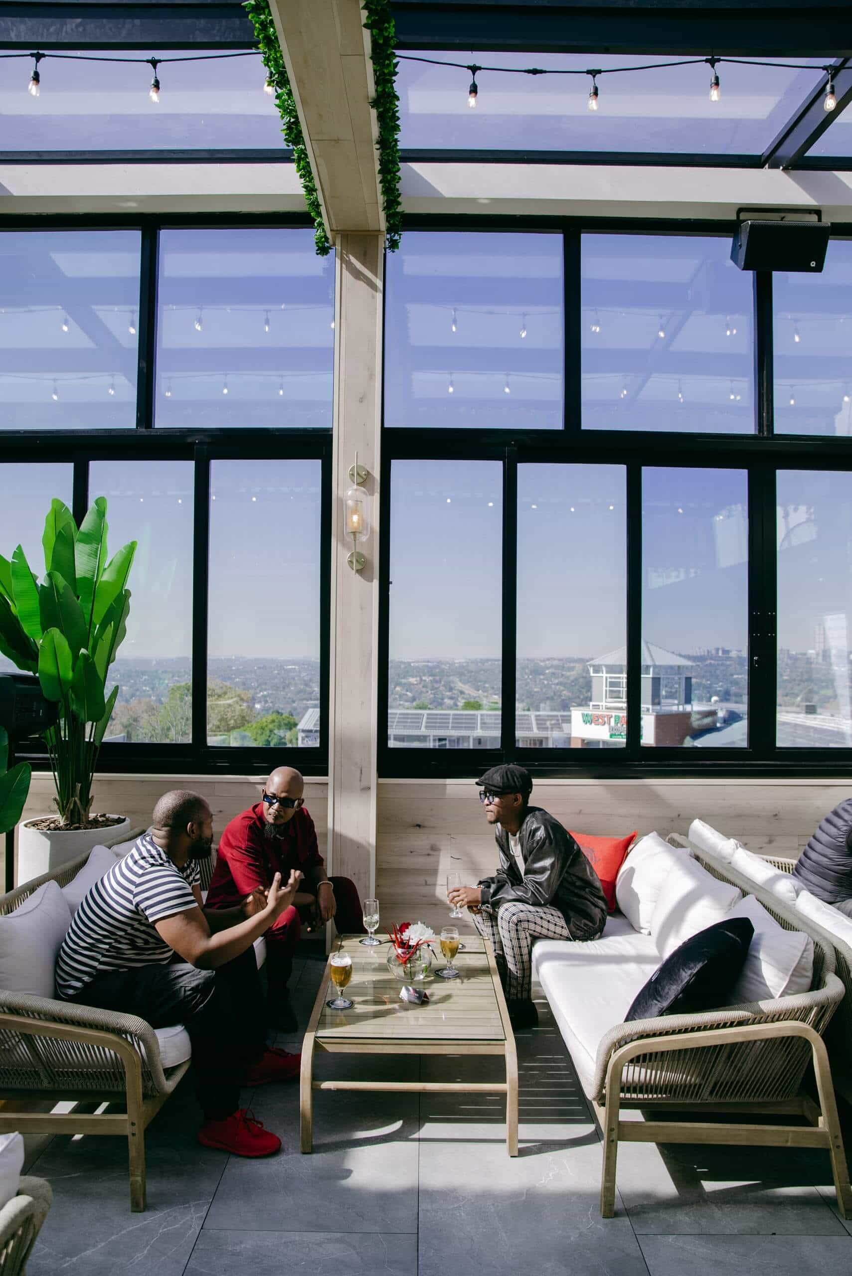 Three men sit and converse around a glass coffee table on a modern rooftop lounge with large windows, cushioned seating, potted plants, and city views in the background under a clear blue sky. Drinks and snacks are on the table.