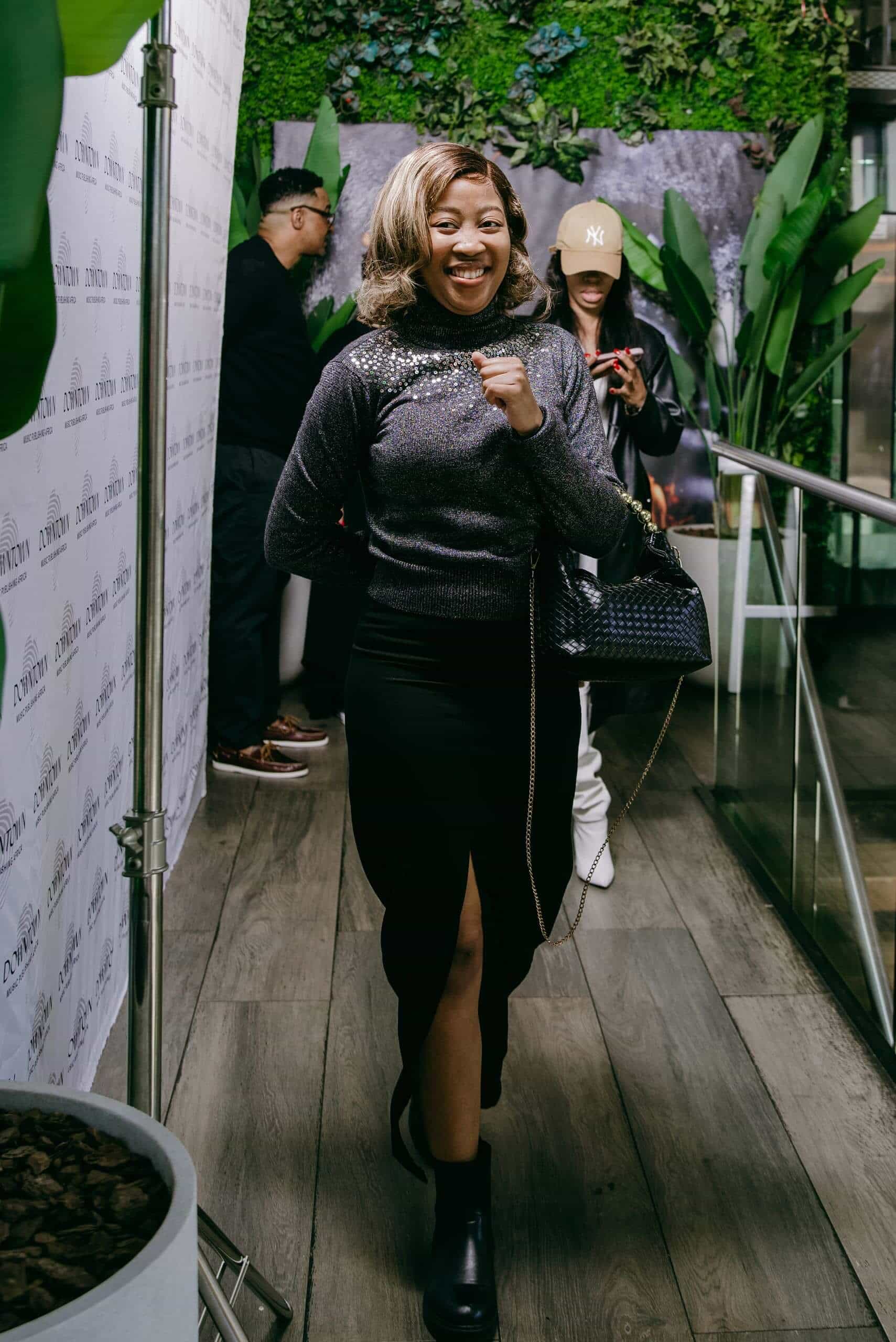 A smiling woman in a sparkly black top, black skirt with a front slit, and ankle boots confidently walks indoors on a wooden floor. Behind her, two people stand near a green wall; one wears a cap and holds a phone, the other faces away.