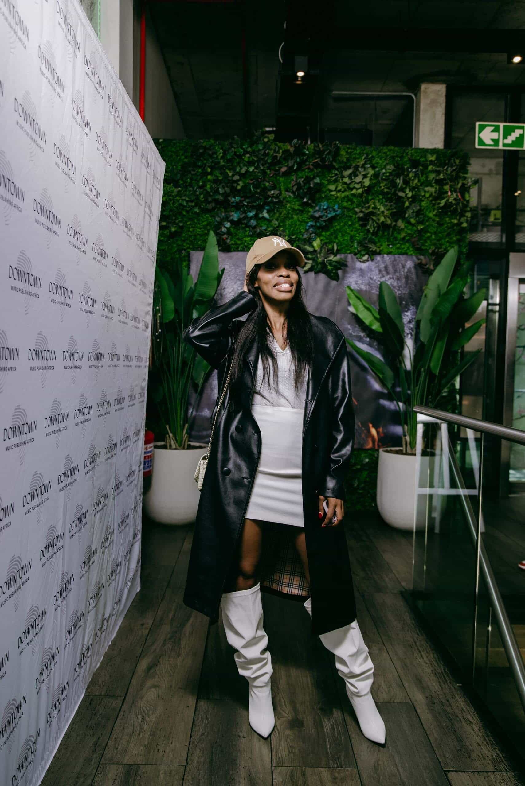 A woman stands indoors smiling, wearing a beige cap, long black leather coat, white dress, and tall white boots. She poses near a Downtown-branded backdrop and lush green plants, with one hand holding her cap. The setting has wooden floors and glass railings.