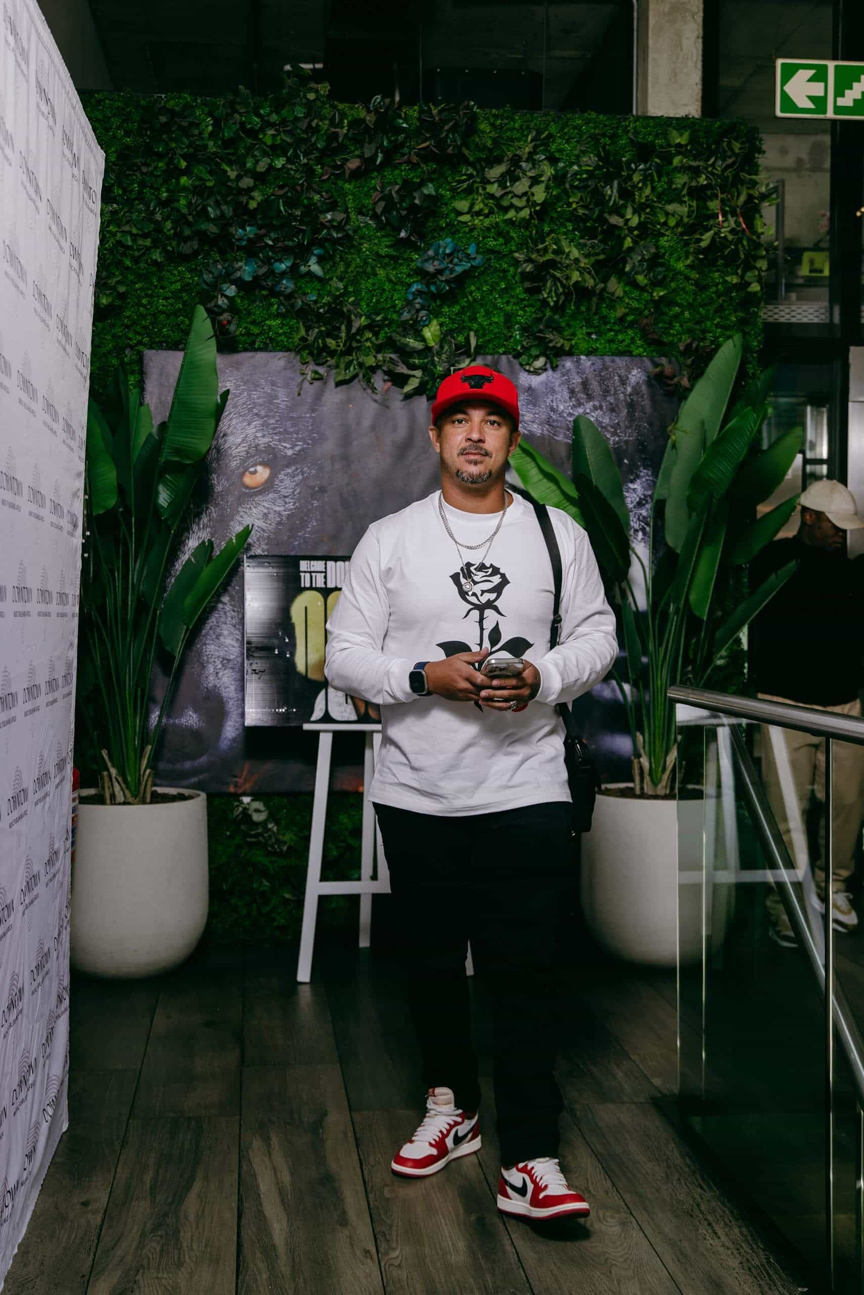 A man stands indoors on a wooden floor, in front of a green plant wall and a dark poster. He wears a white long-sleeve shirt, black pants, red and white sneakers, and a red cap. He holds a drink and has sunglasses on his cap. Two large potted plants frame him.