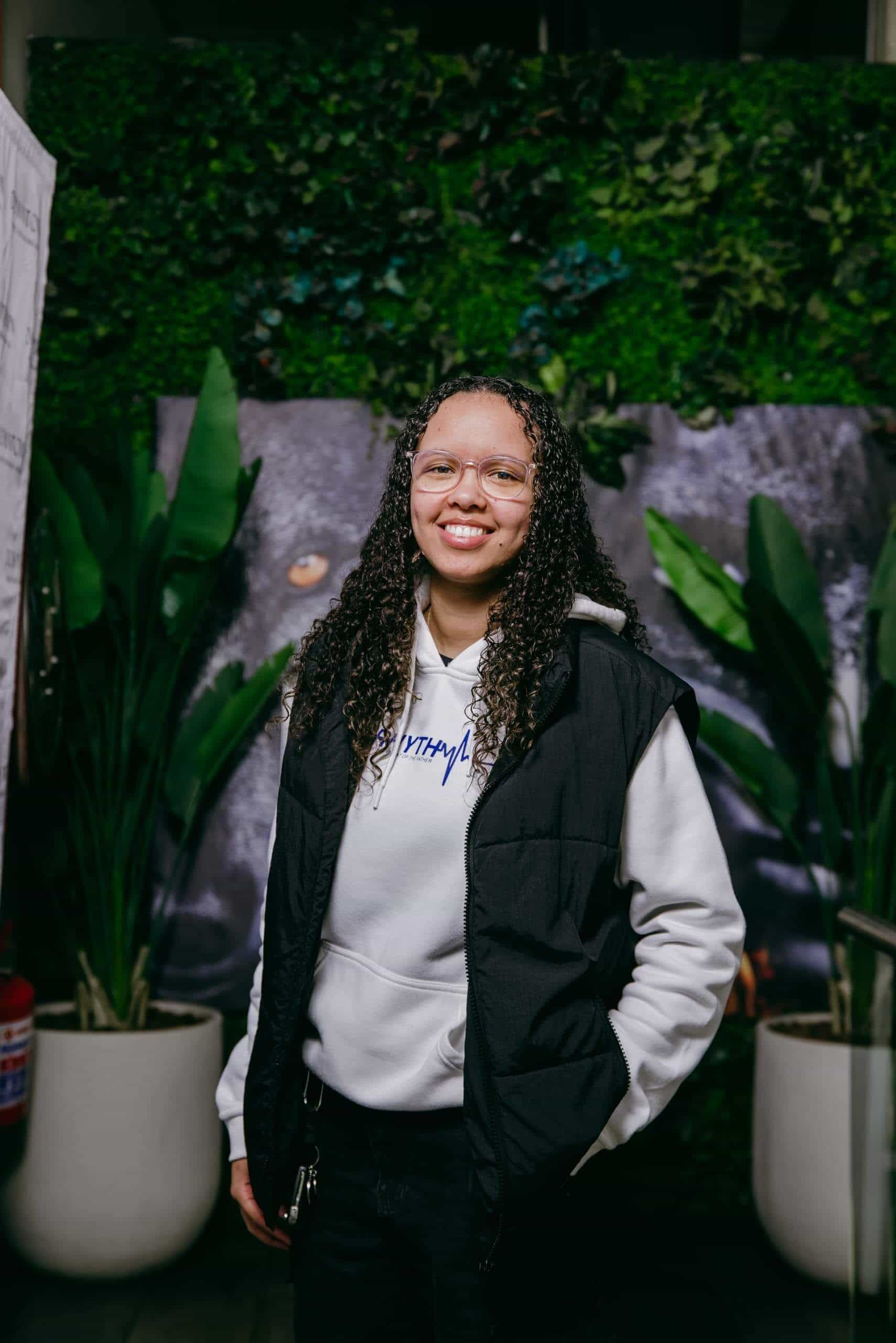 A person with curly hair, glasses, and a friendly smile stands indoors wearing a white hoodie and black vest. They have one hand in their pocket. Behind them are large green plants in white pots and a wall covered in green foliage.