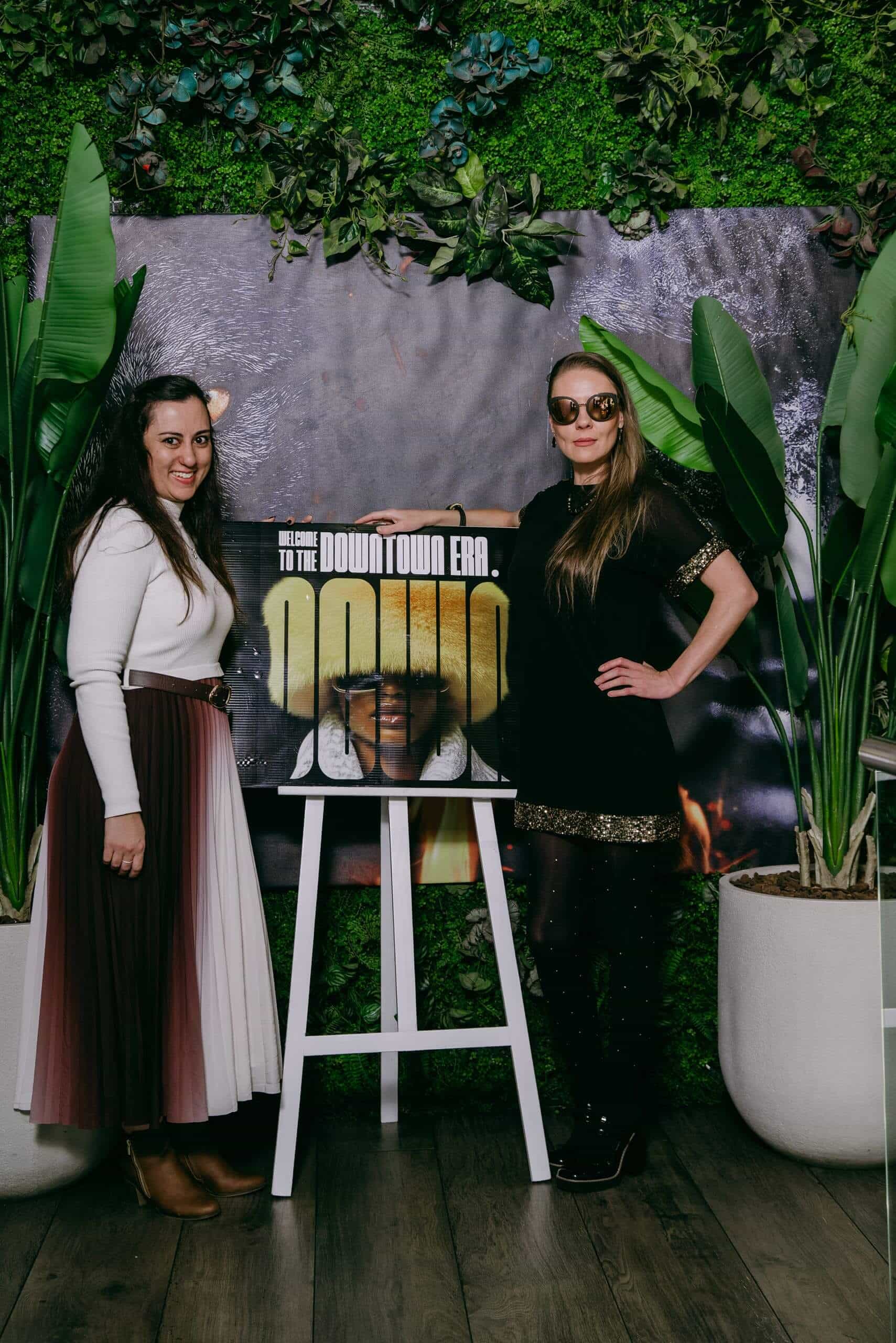 Two women stand on either side of an easel displaying a &ldquo;Welcome to the Downtown Era&rdquo; sign. Both women smile at the camera; one wears a white top with a color-blocked skirt, the other wears a black dress and sunglasses. Large potted plants flank the scene.