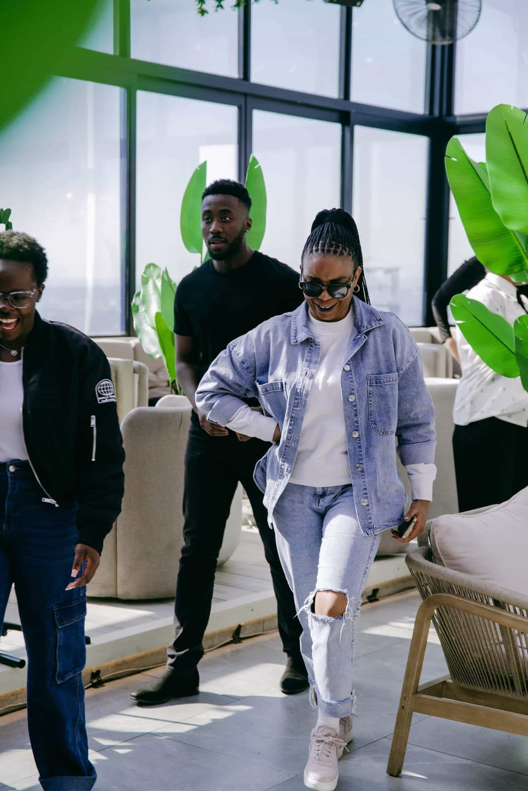Three people are indoors near large windows and green plants. A woman in ripped jeans, a denim jacket, and sunglasses walks and smiles. Another woman in a black jacket and glasses is laughing. A man in black stands behind them. The setting appears bright and modern.